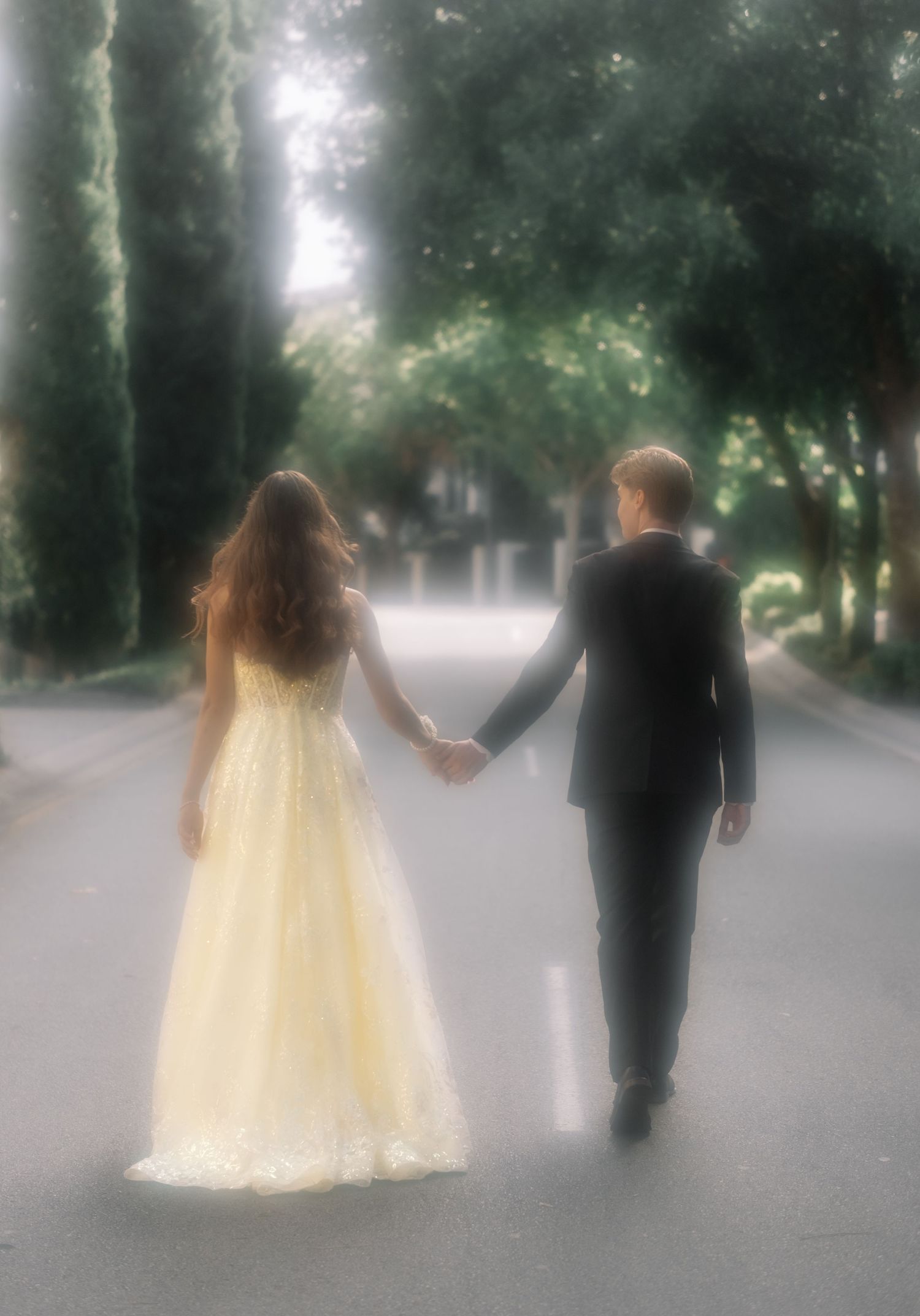 Couple holding hands walking down a tree-lined path, woman in yellow lace gown and man in suit.