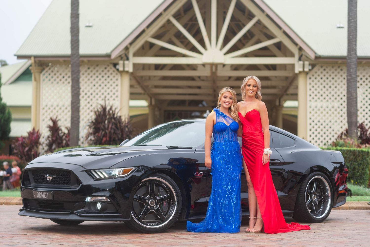 Two women in blue and red formal gowns posing beside black Ford Mustang at prom.