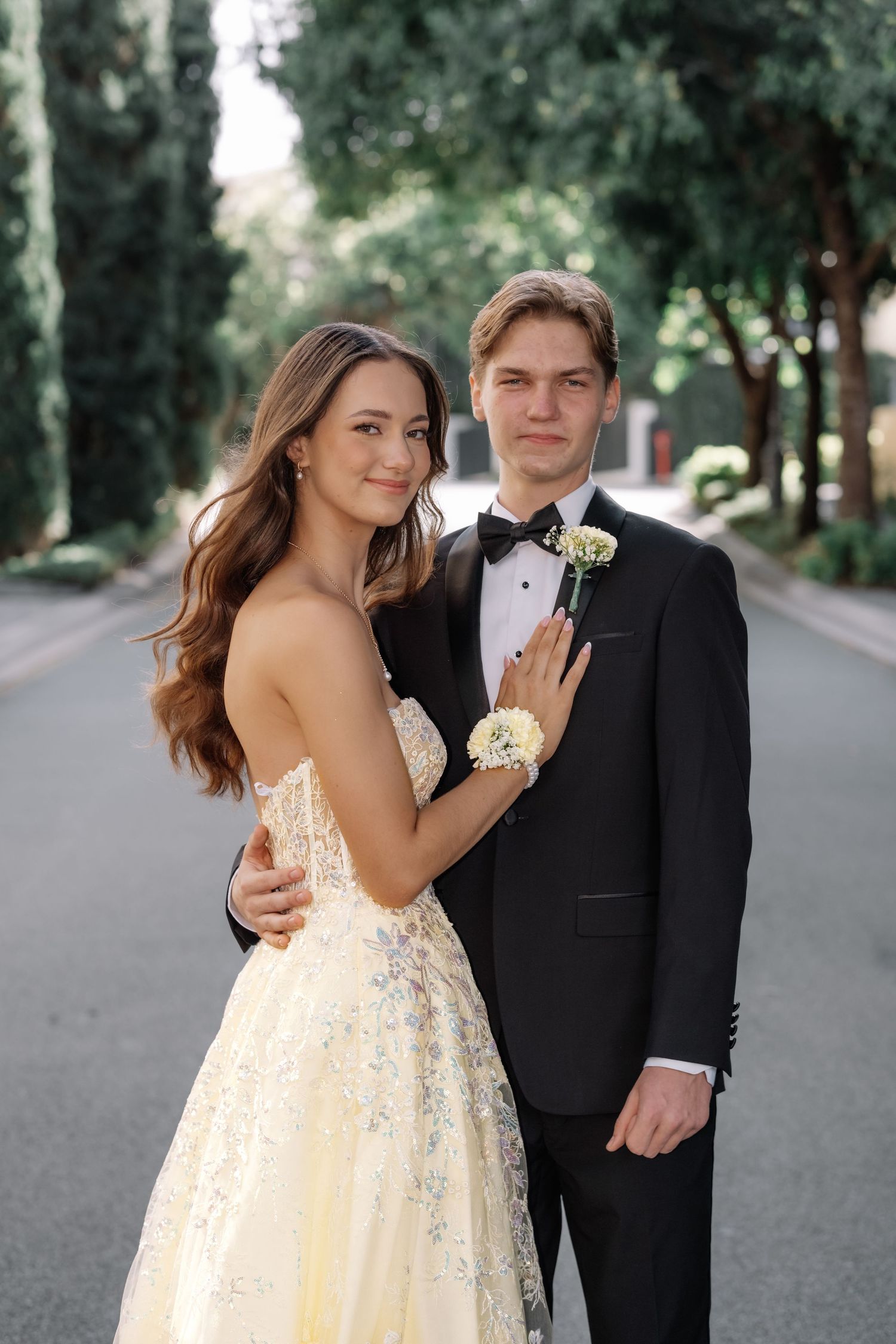 Prom couple in formal attire posing on tree-lined street, woman in yellow ball gown.