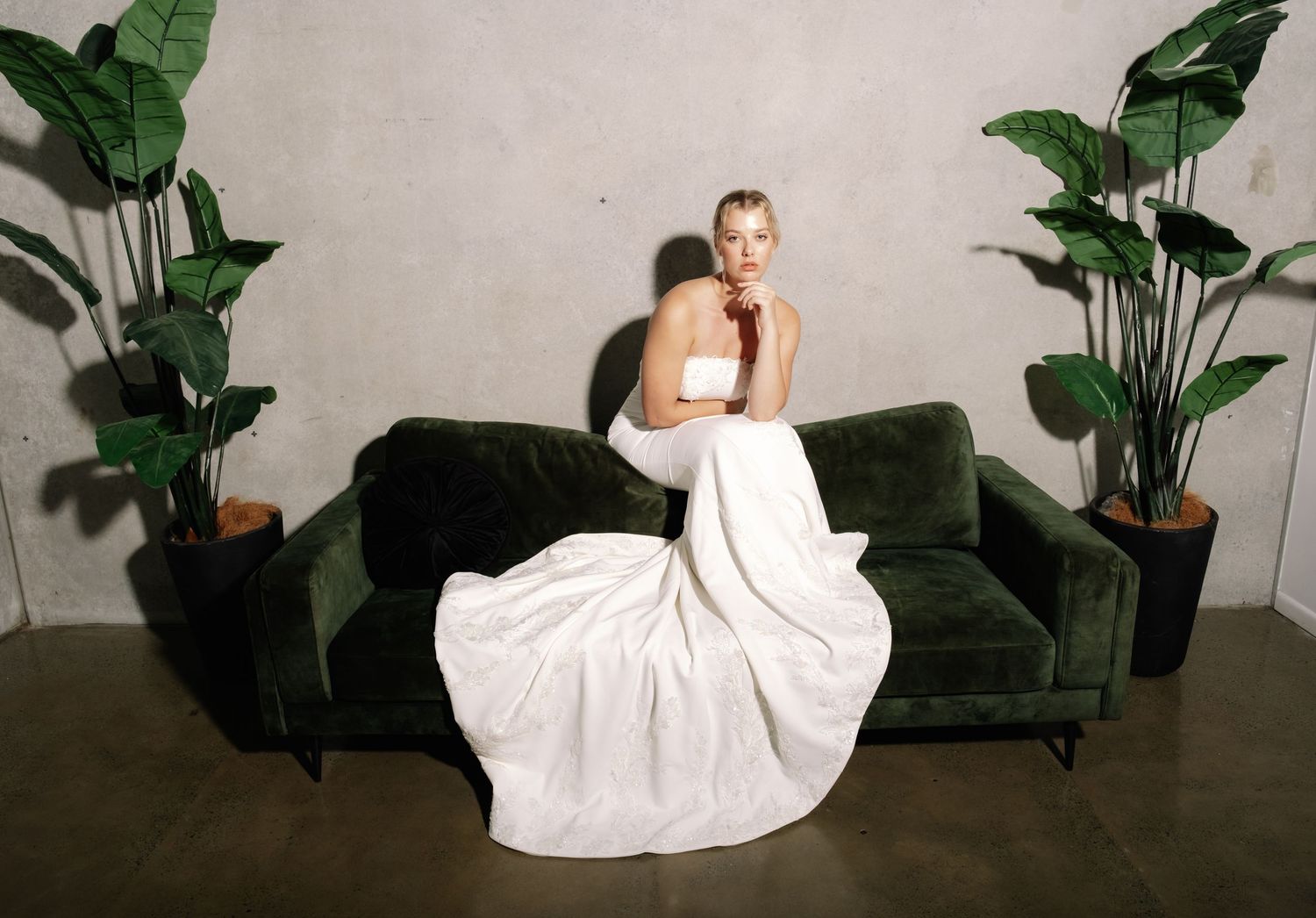Blonde bride in minimalist strapless white gown sits atop green velvet sofa flanked by tropical plants.