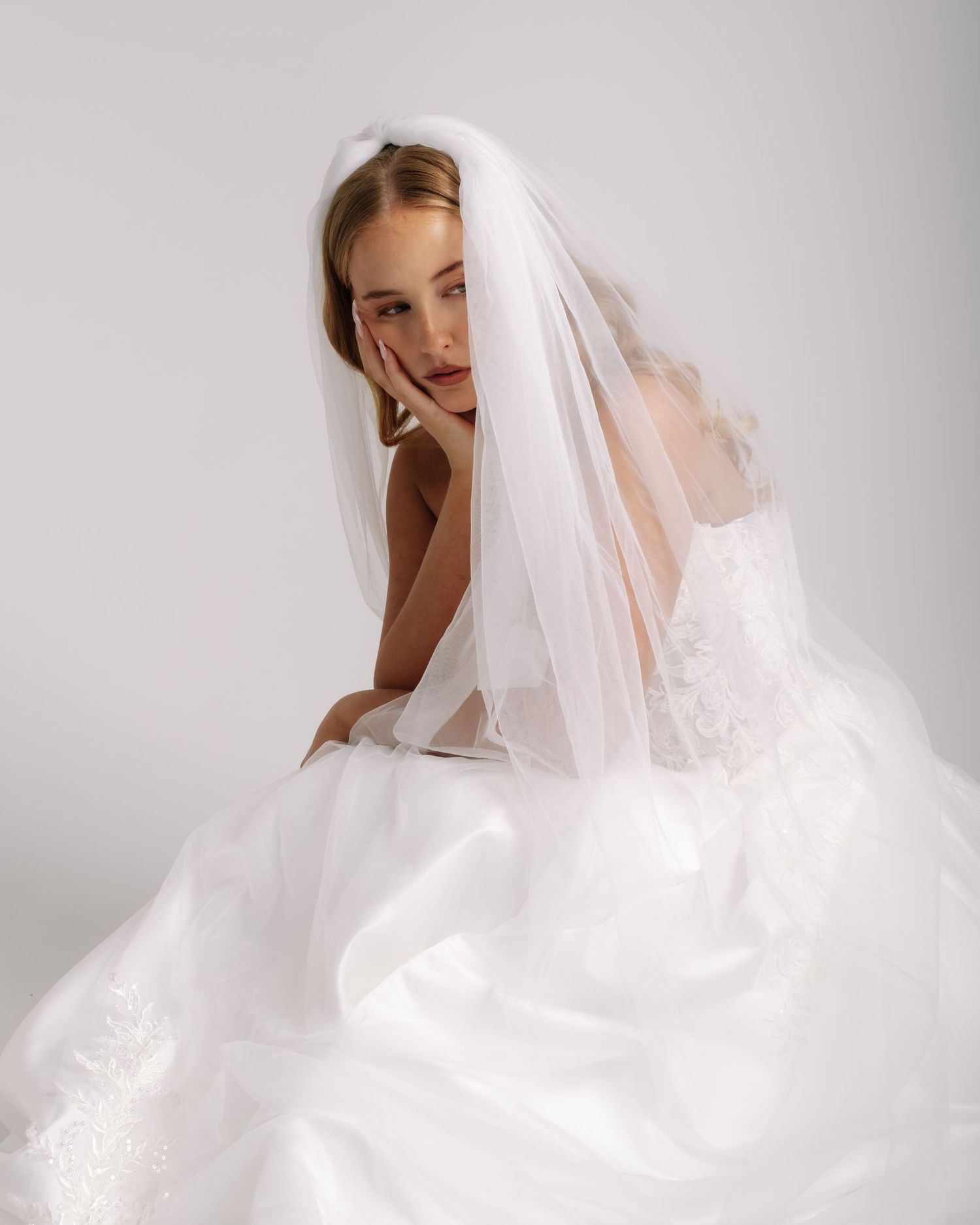 Elegant bride in a white lace wedding gown and veil posing gracefully on a white background.