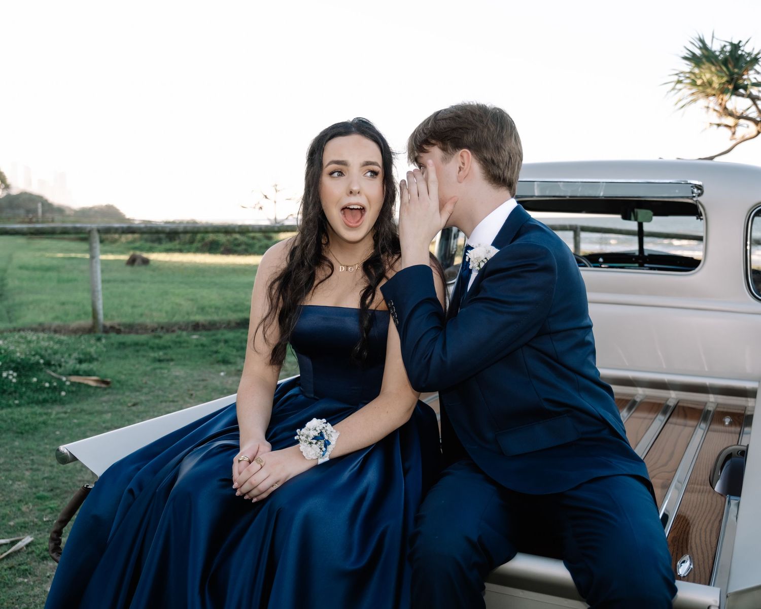 Teenage couple in formal prom attire sitting on a vintage car, boy whispering to girl.