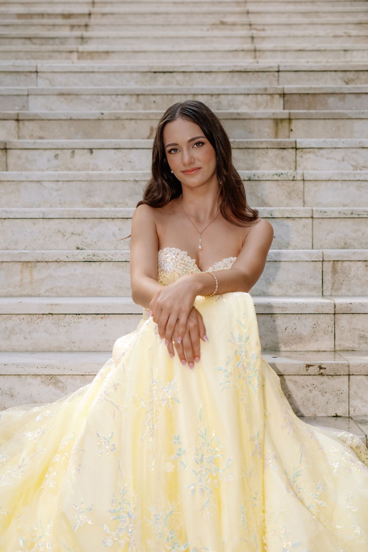 Young woman in yellow floral ball gown sitting on marble steps, wearing pearl jewelry.