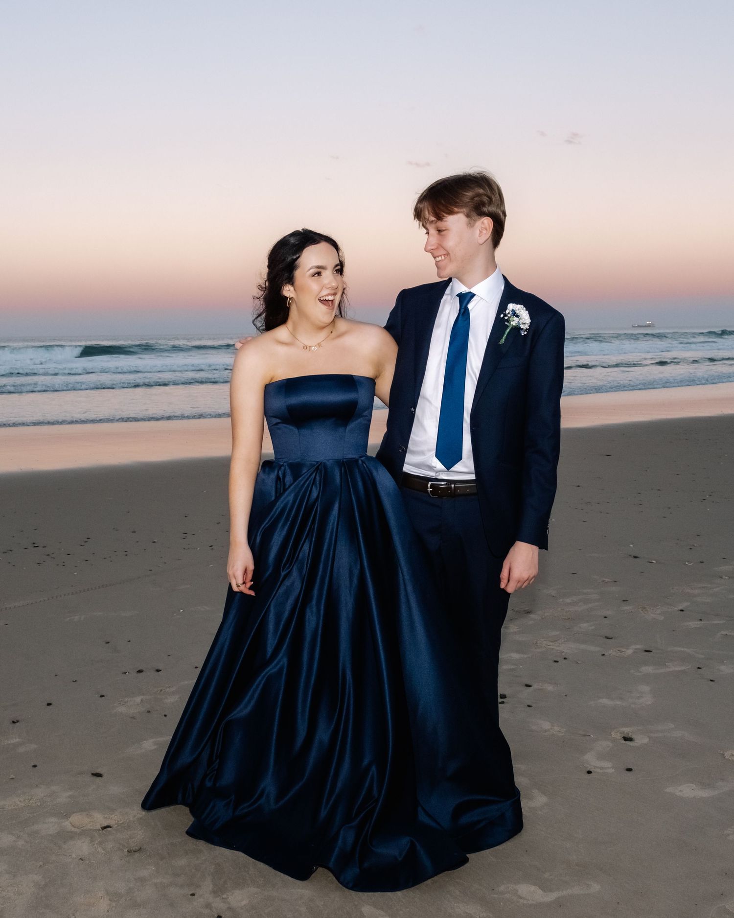 Couple in formal wear, navy ballgown and navy suit, smiling on a beach at sunset.