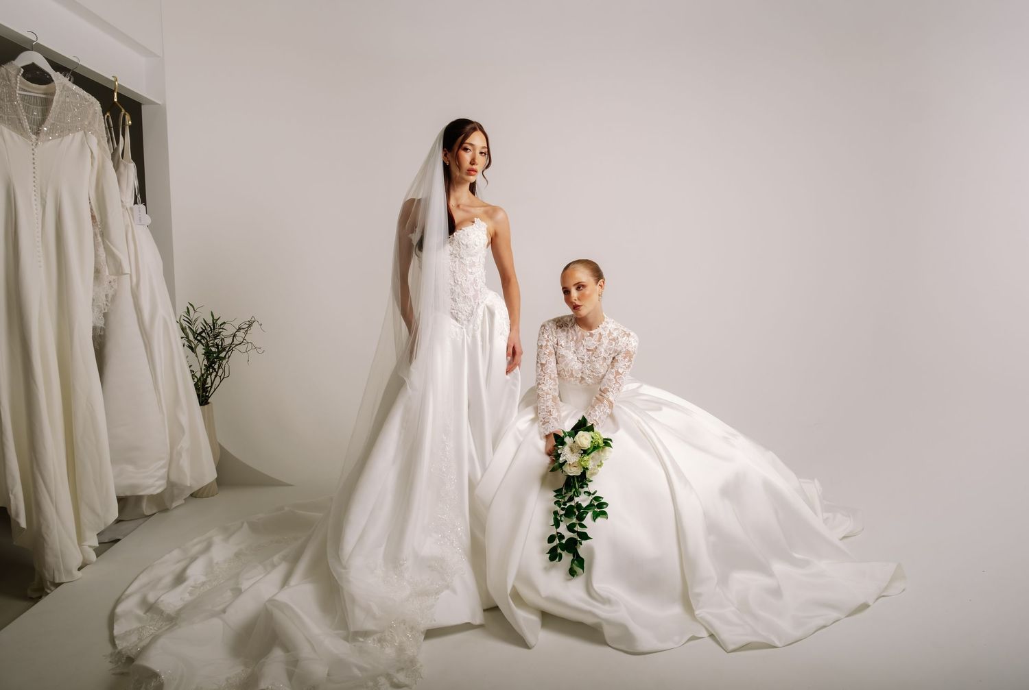 Two brides in elegant white wedding dresses pose gracefully in a bright bridal boutique studio.