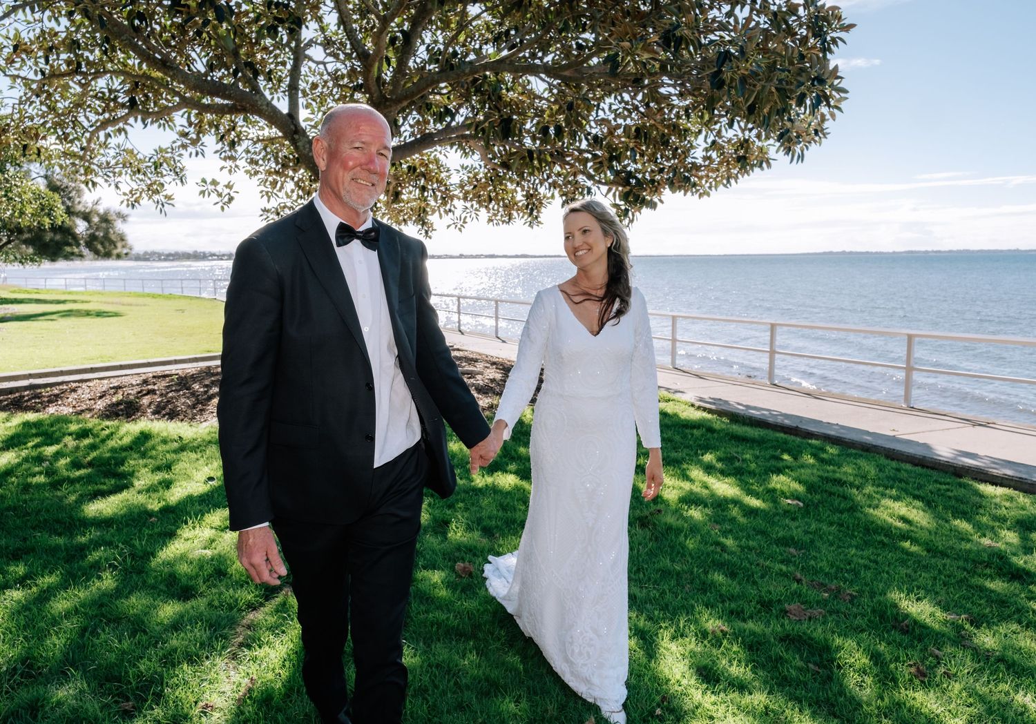 Bride and groom holding hands walking on green lawn with scenic ocean waterfront backdrop.