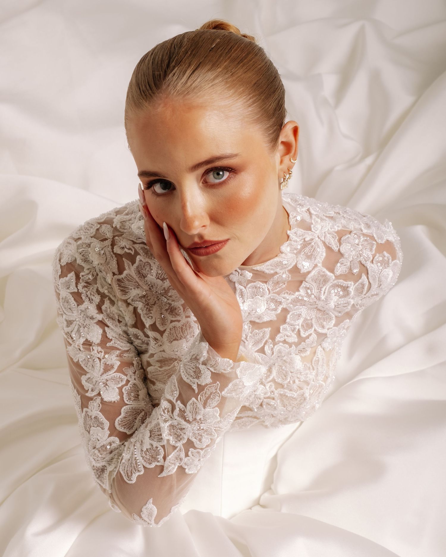Redhead bride in sheer floral lace long-sleeve gown rests cheek on hand while lying on white satin.