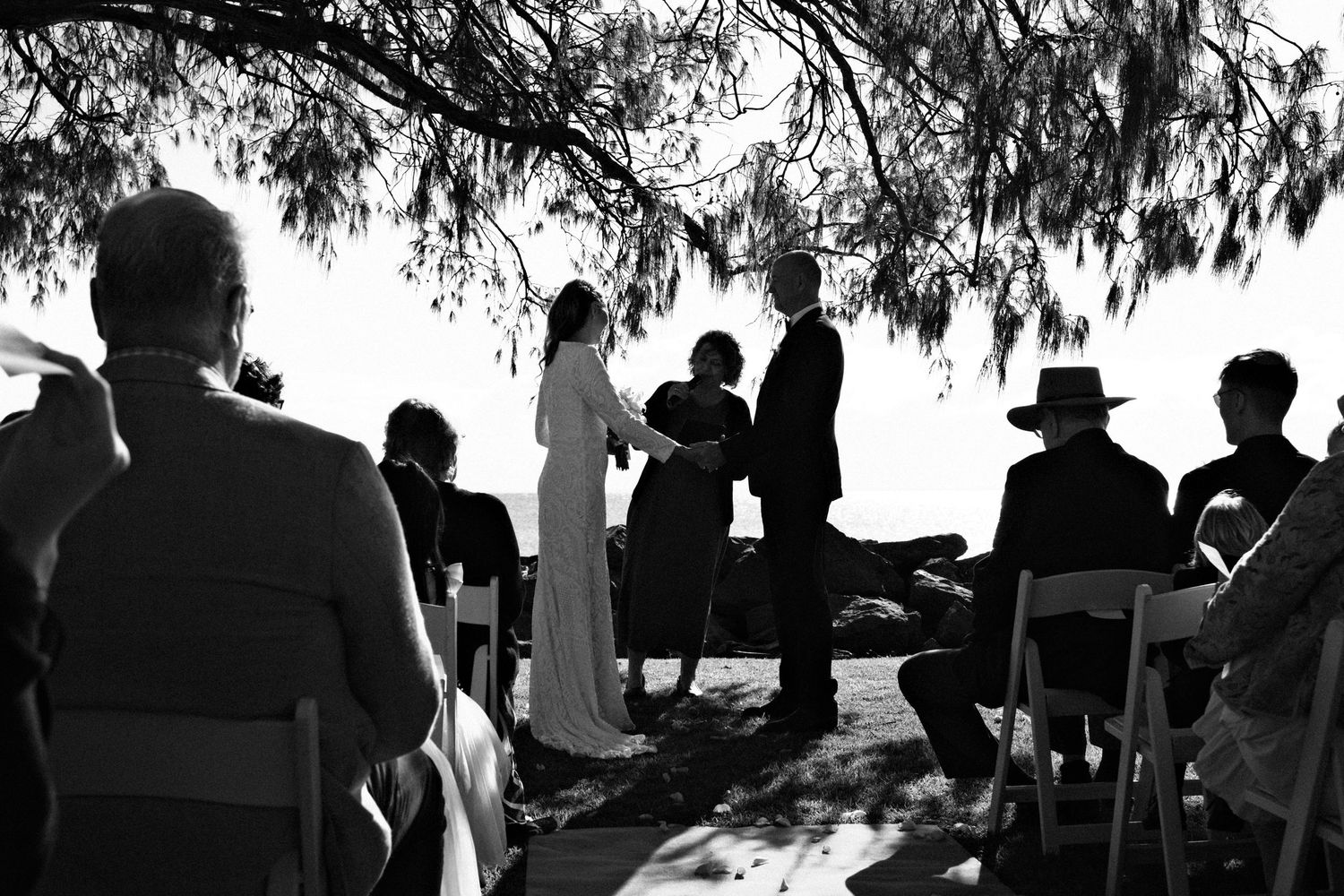 Black and white silhouette of a wedding ceremony with guests seated under a tree outdoors.