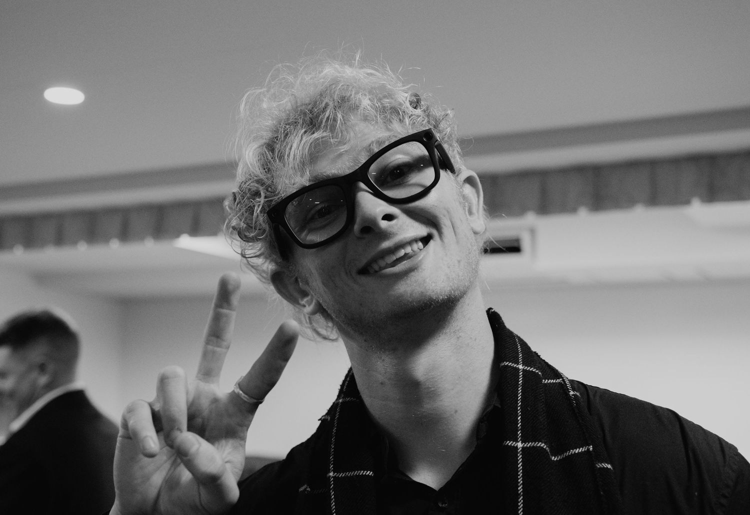 Young curly-haired man with glasses making peace sign in black and white photo.