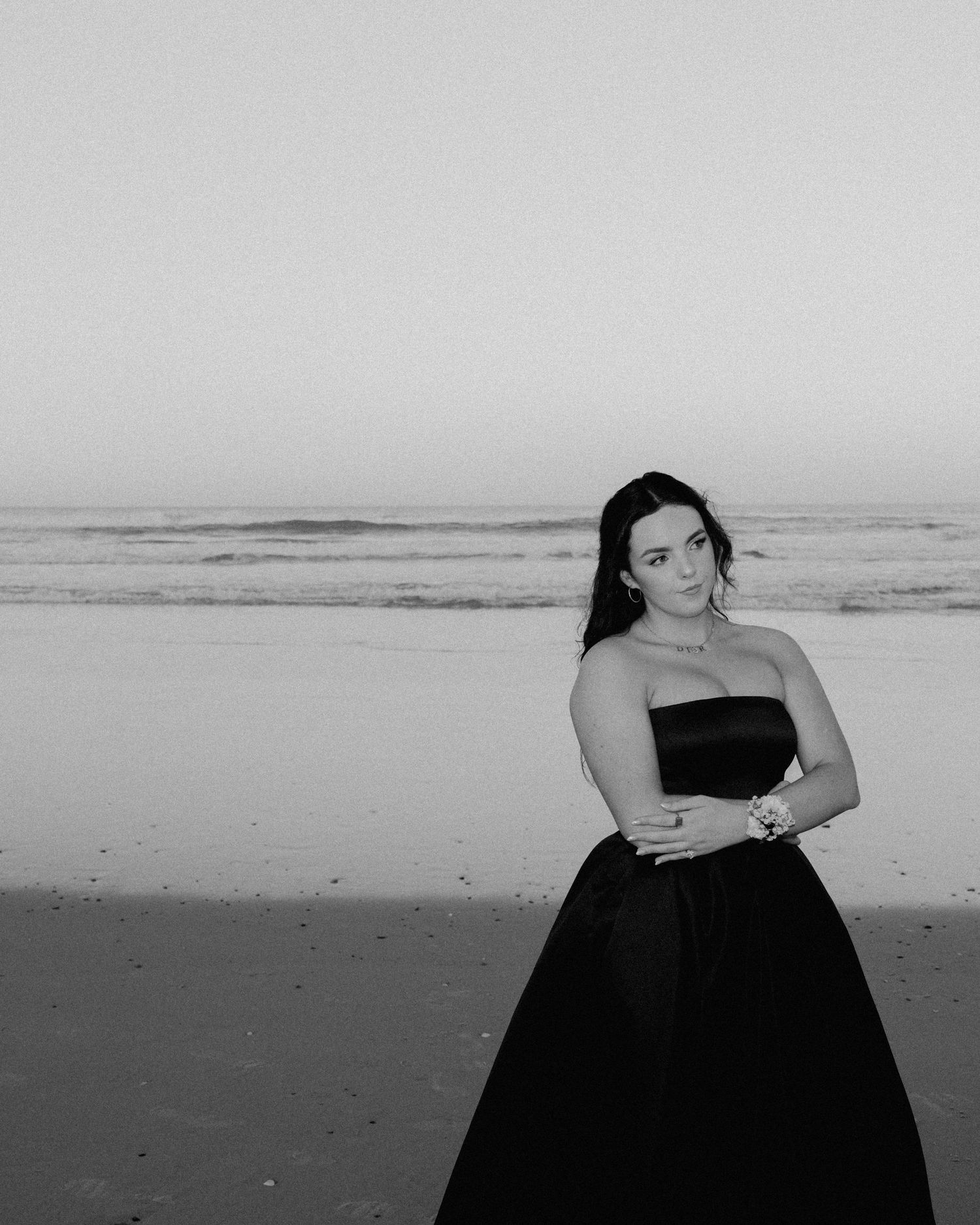 Black and white photo of a woman in a strapless ball gown posing elegantly on a beach at dusk.