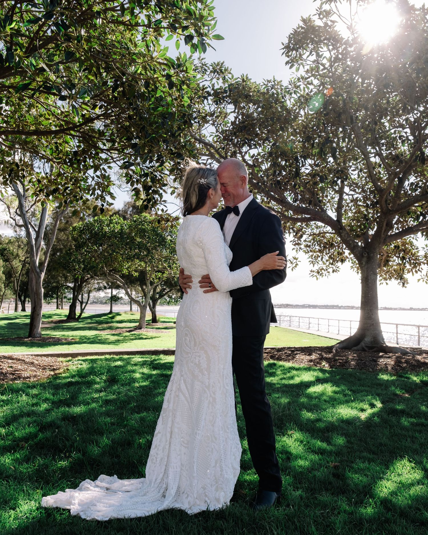 Bride and groom sharing a romantic embrace under trees with ocean views at sunset.