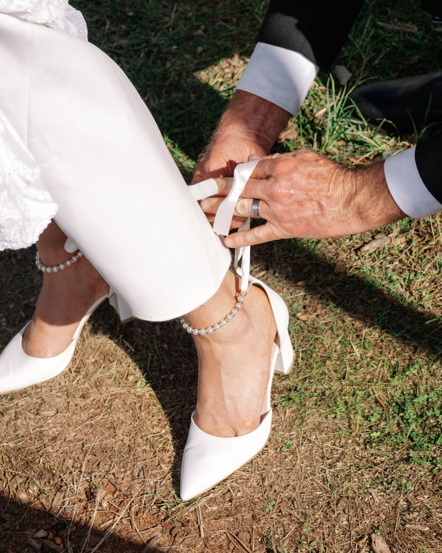 Groom carefully tying ribbon on bride's white pointed-toe heels with pearl anklet detail.
