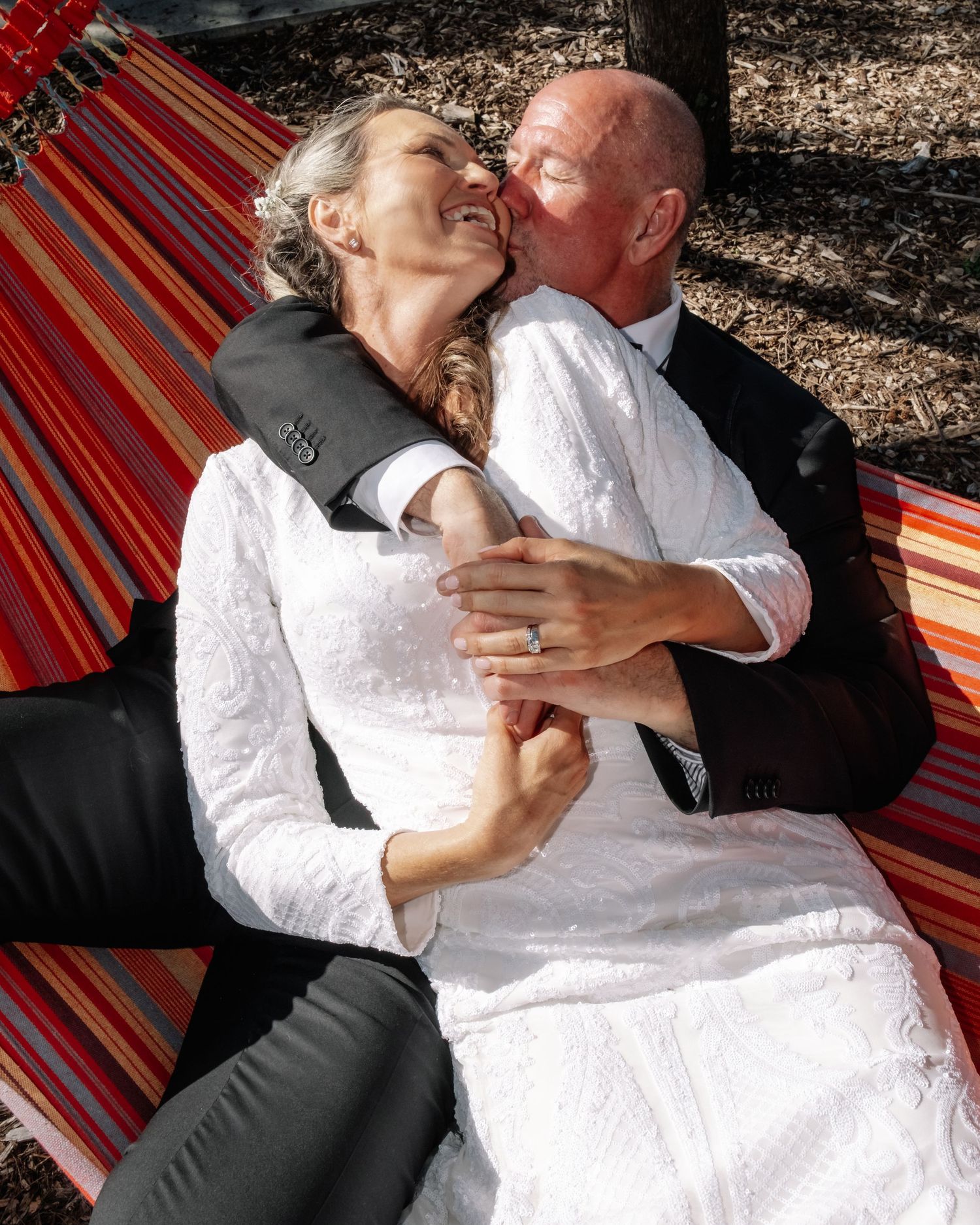 Older groom kissing bride on cheek while relaxing in striped hammock on wedding day.