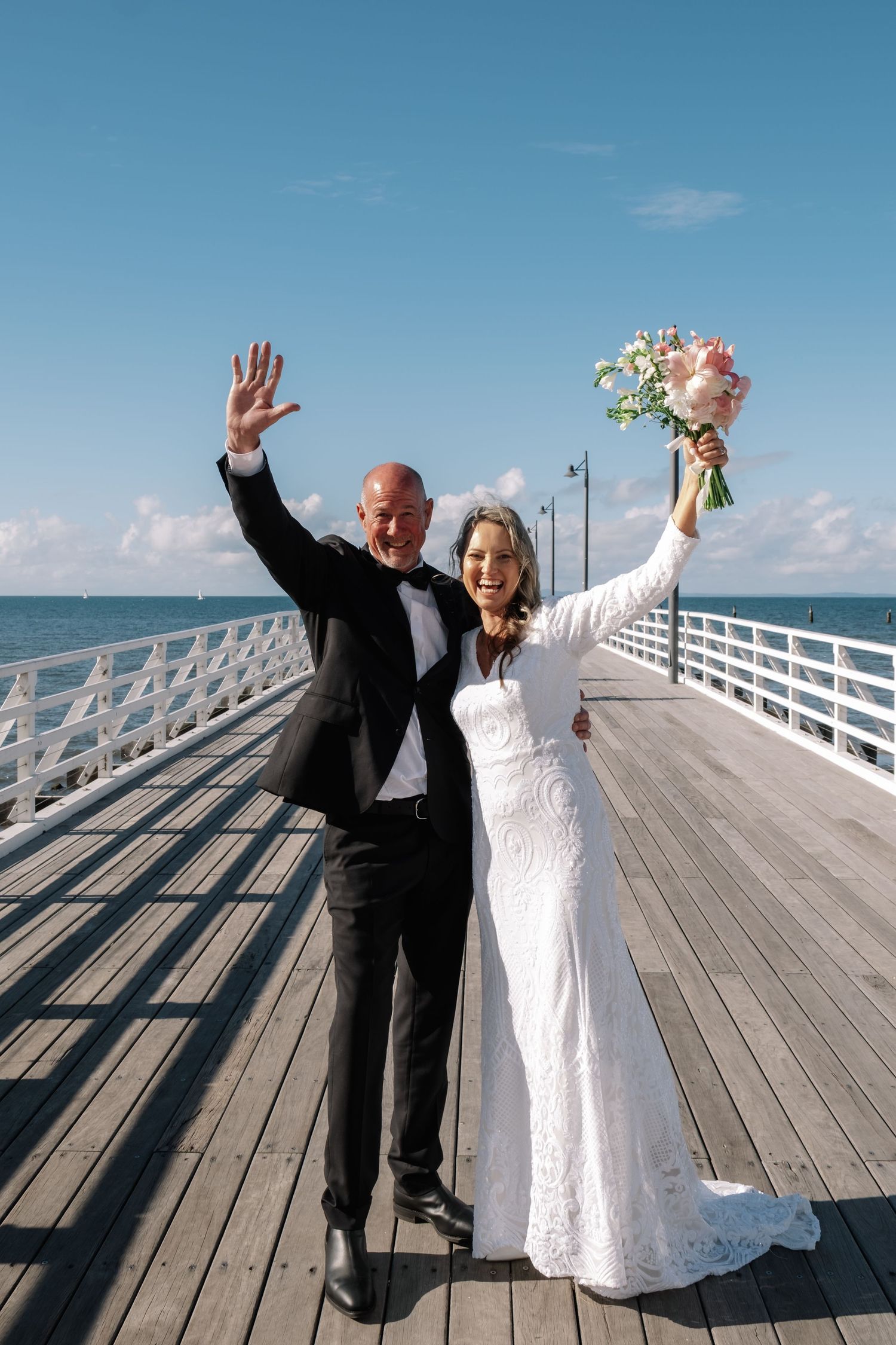 Happy newlywed couple raising their arms in celebration on a wooden pier overlooking the ocean.