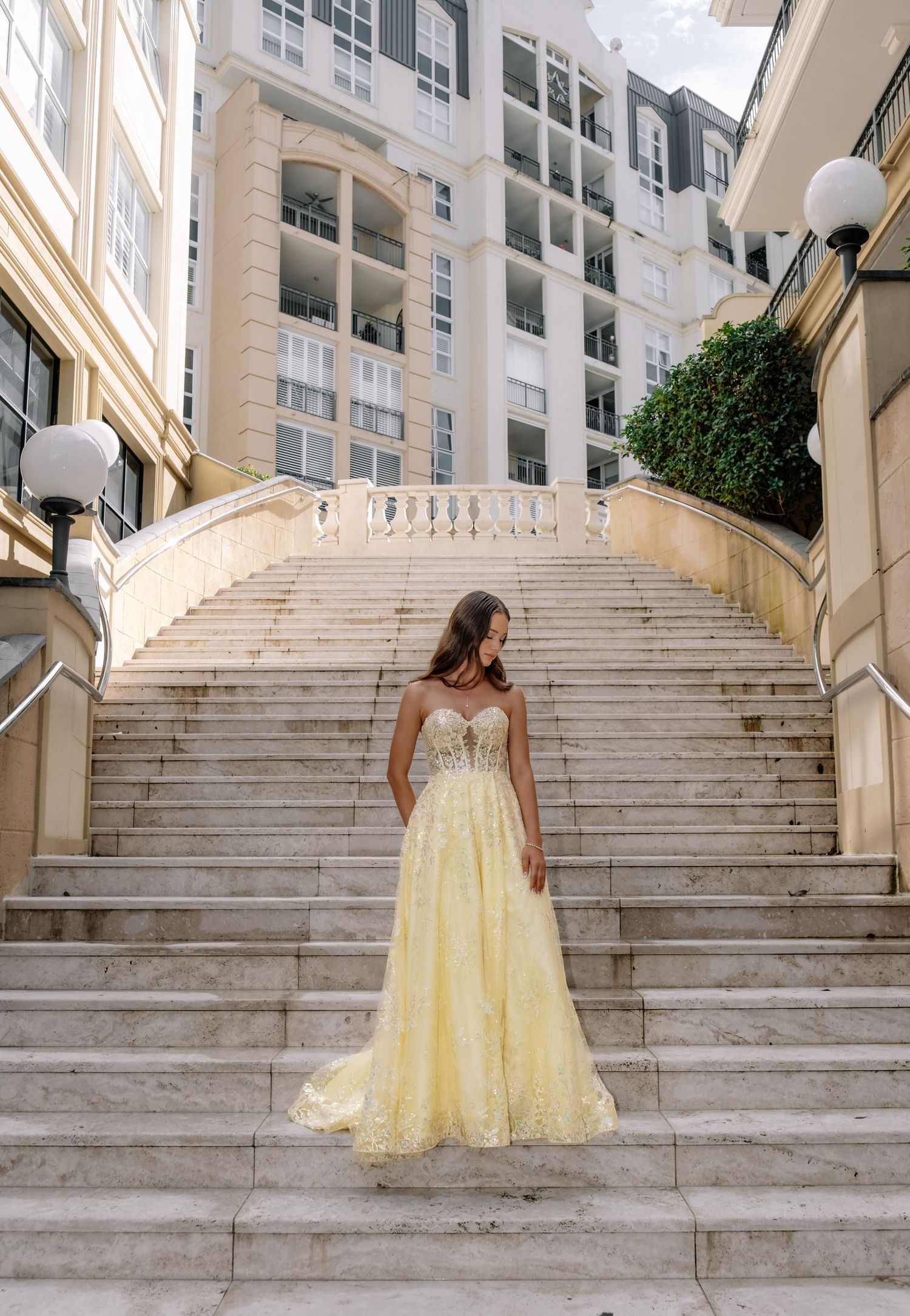 Elegant woman in yellow lace ball gown standing on grand marble staircase outdoors.