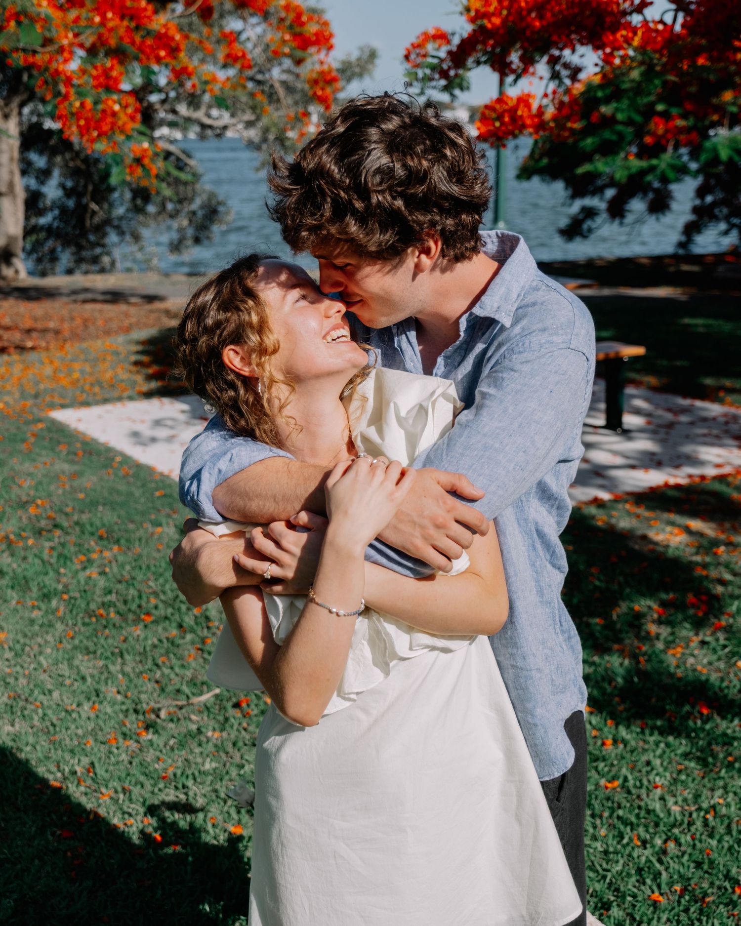 Joyful couple embracing and laughing under red flowering trees during an engagement shoot.