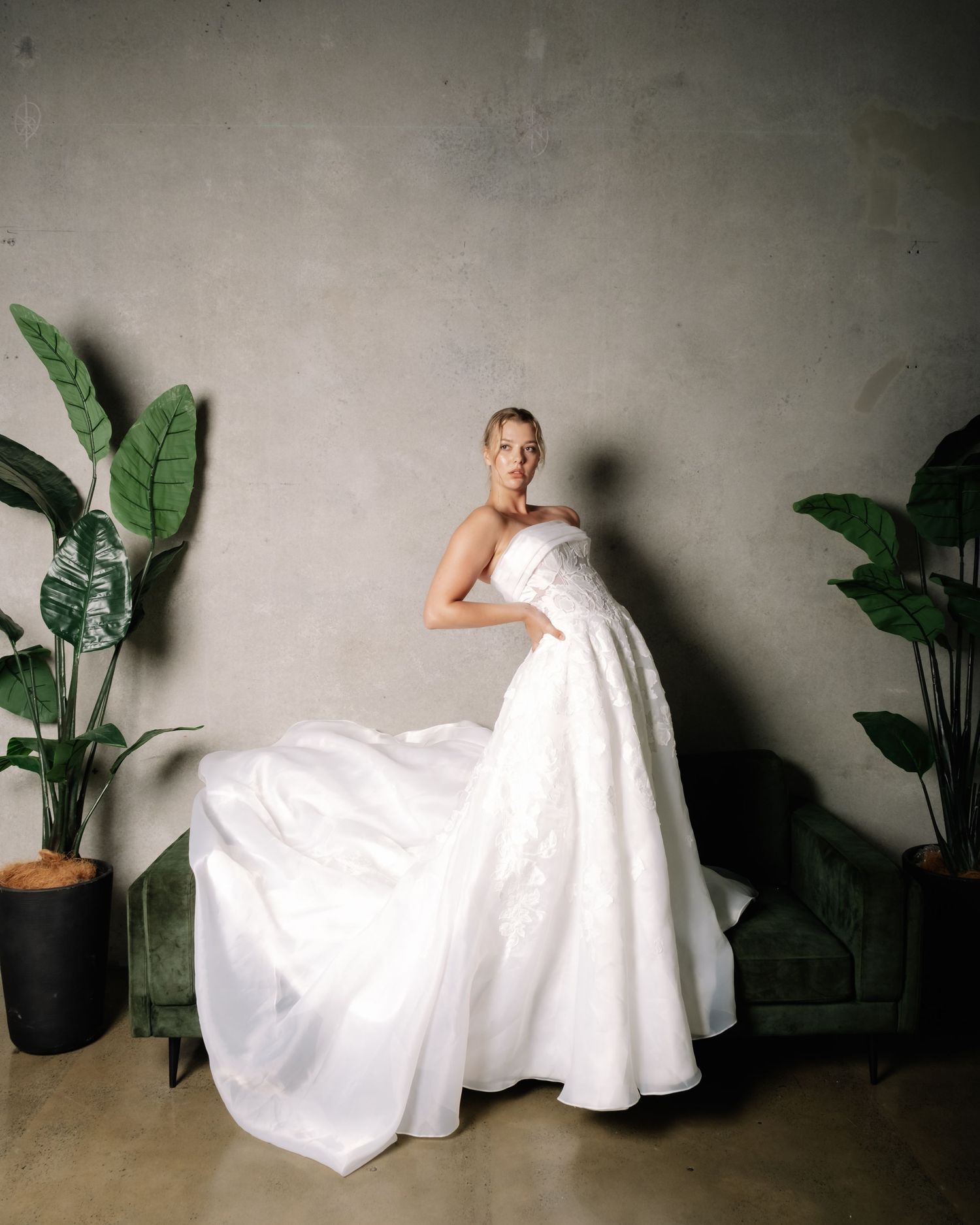 Bride twirls strapless white ballgown with flowing train beside green velvet sofa and large tropical plants.