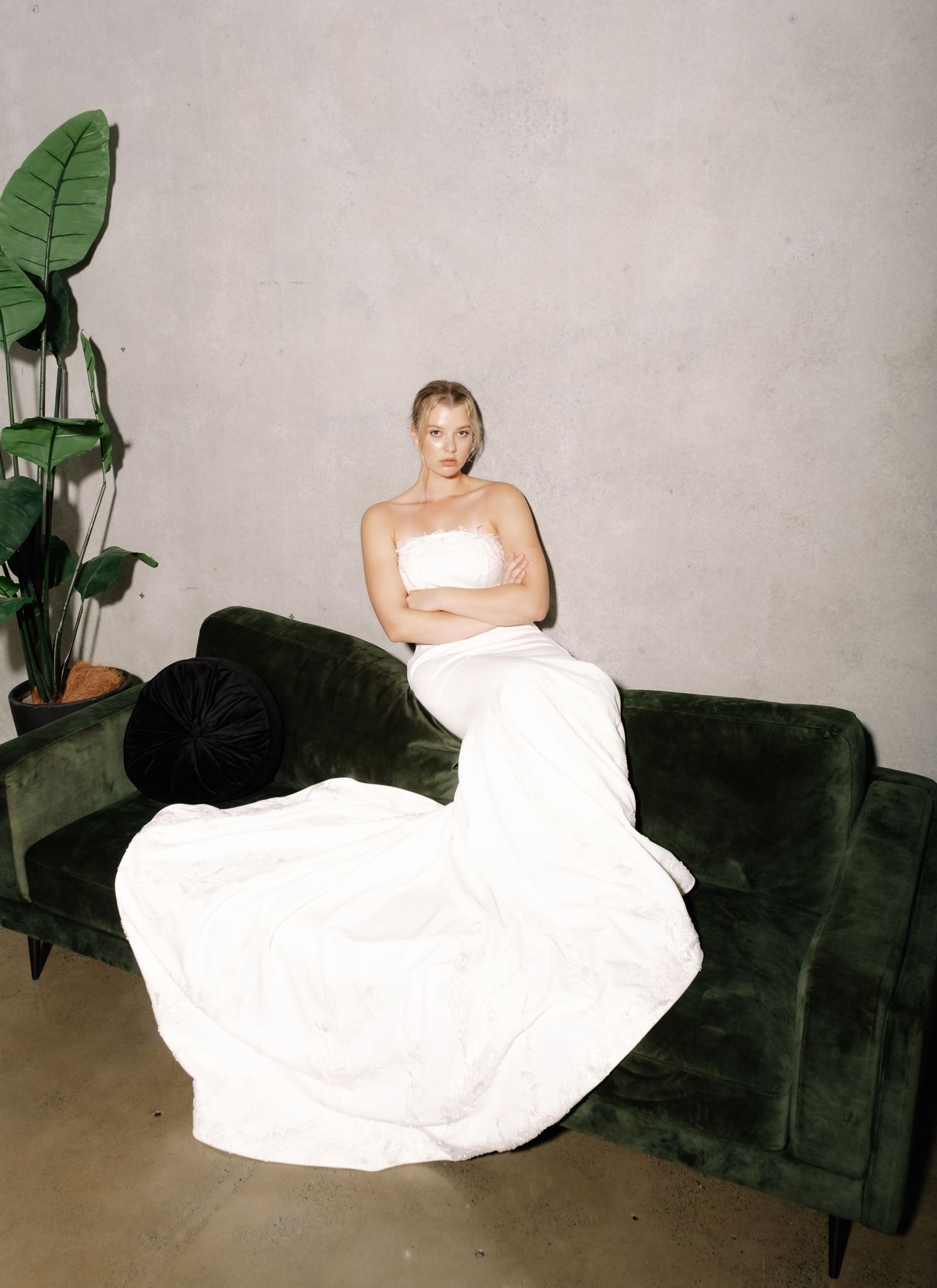 Blonde woman in a white strapless gown sitting on a green velvet sofa beside a tropical plant.