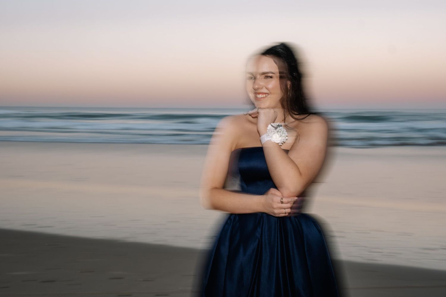 Motion blur photo of a smiling woman in a navy strapless ballgown on a beach at sunset.