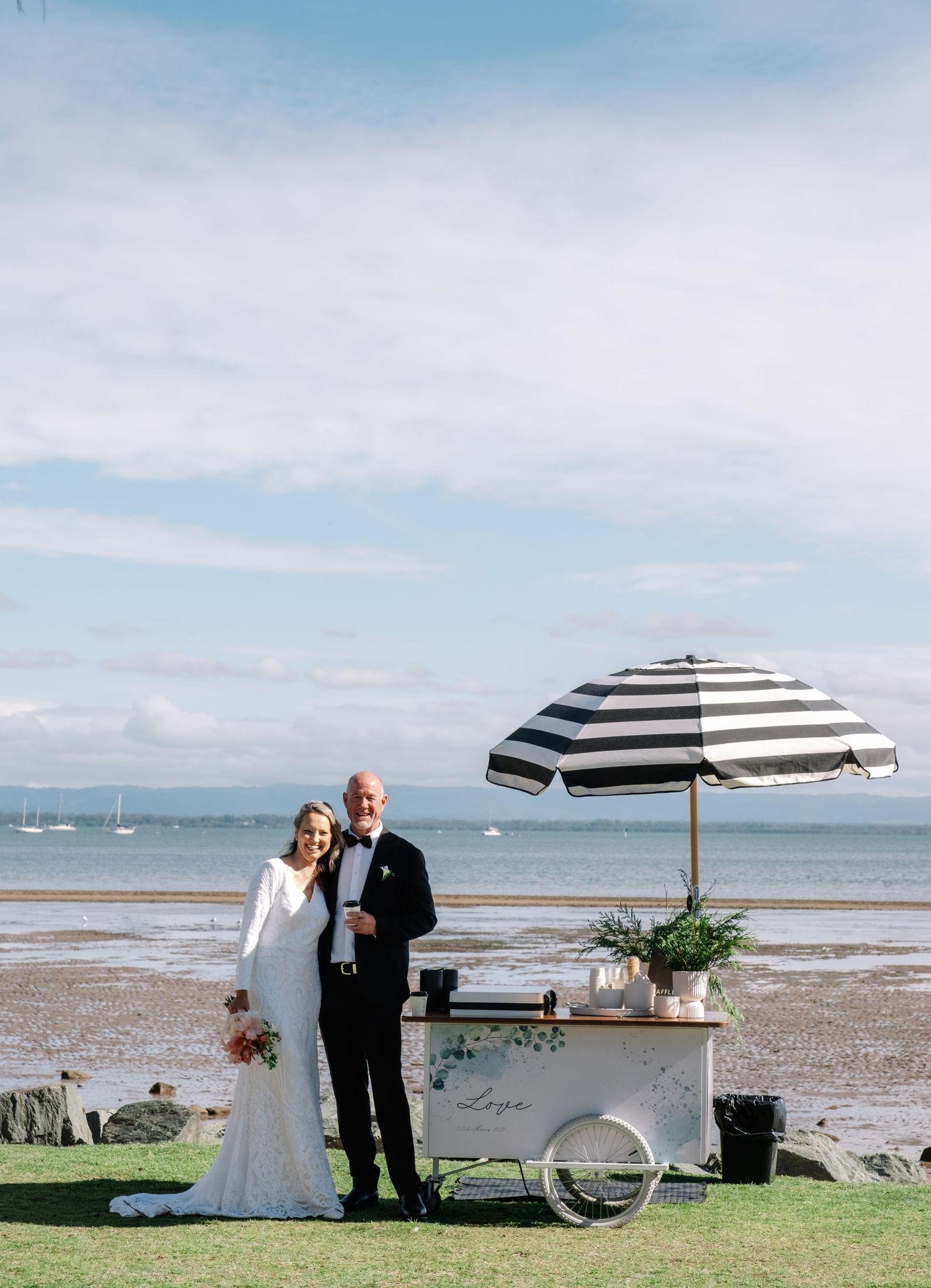 Bride and groom posing beside a vintage ice cream cart at a scenic seaside wedding venue.