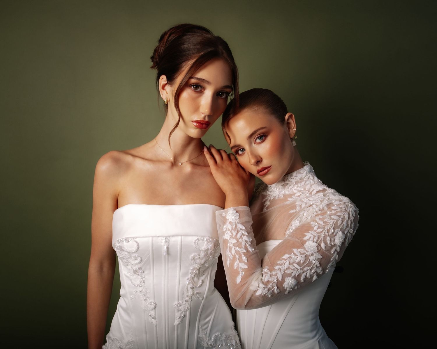 Two brides pose together, one in strapless corset gown, other in long-sleeve floral lace wedding dress.