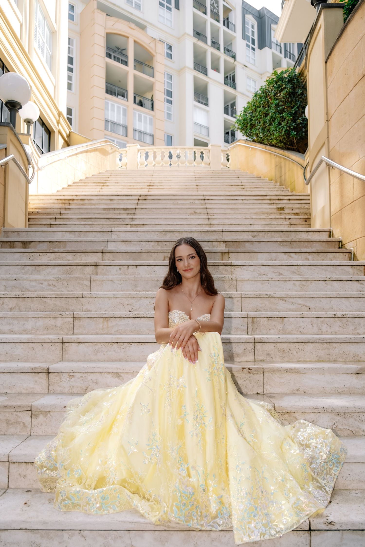 Woman in yellow embroidered strapless ball gown seated on marble steps near building.