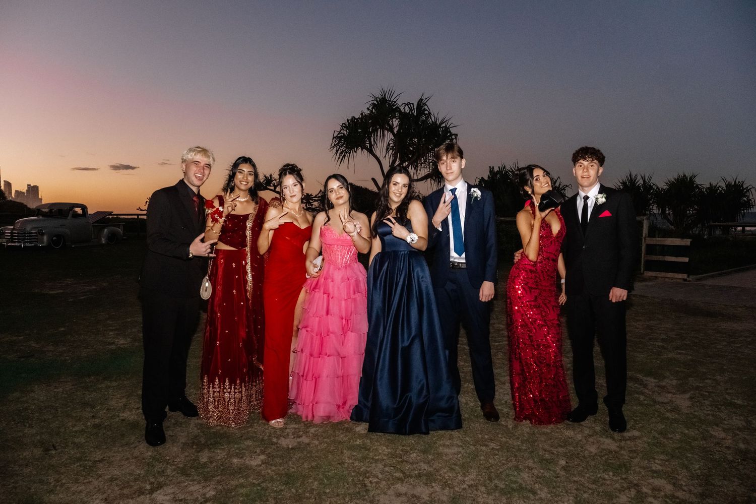 Group of high school students in colorful formal prom attire posing outdoors at sunset.