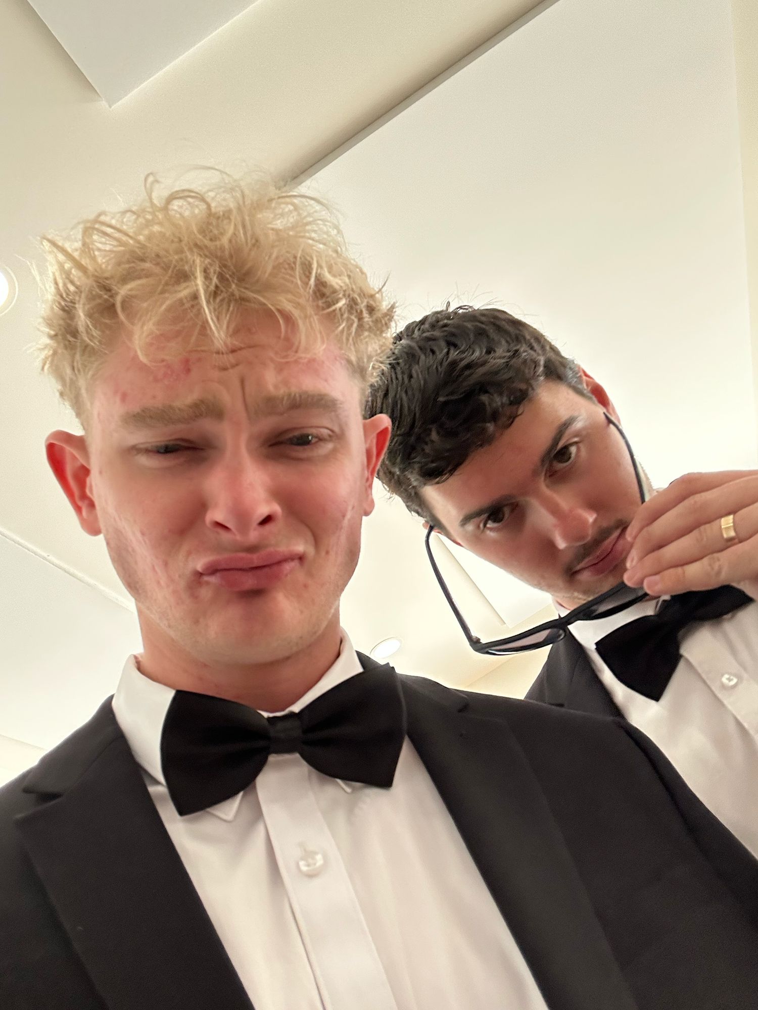 Two young men in black tuxedos with bow ties taking a mirror selfie.