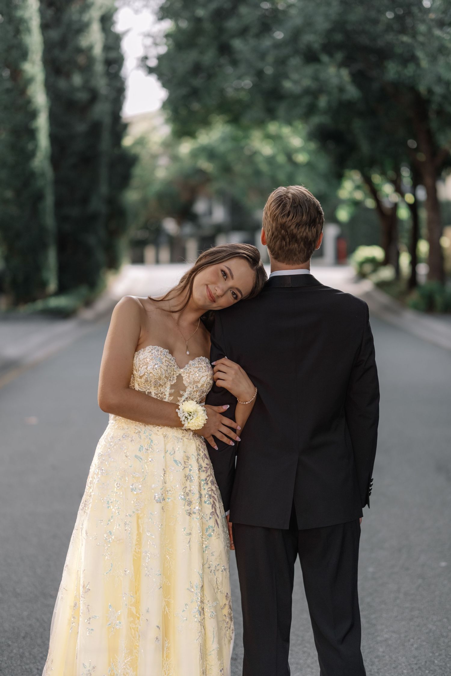 Prom couple posing on tree-lined avenue, woman in yellow gown leaning on mans shoulder.