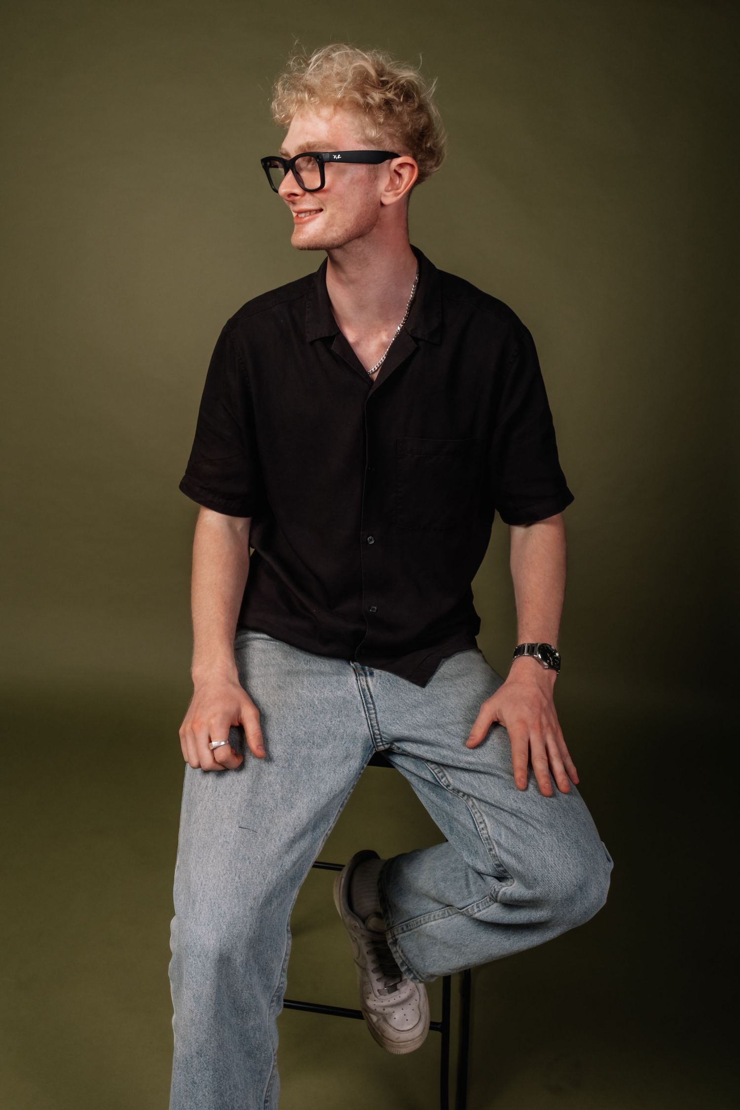 Young blonde man in glasses and casual attire sitting on a stool against green backdrop.