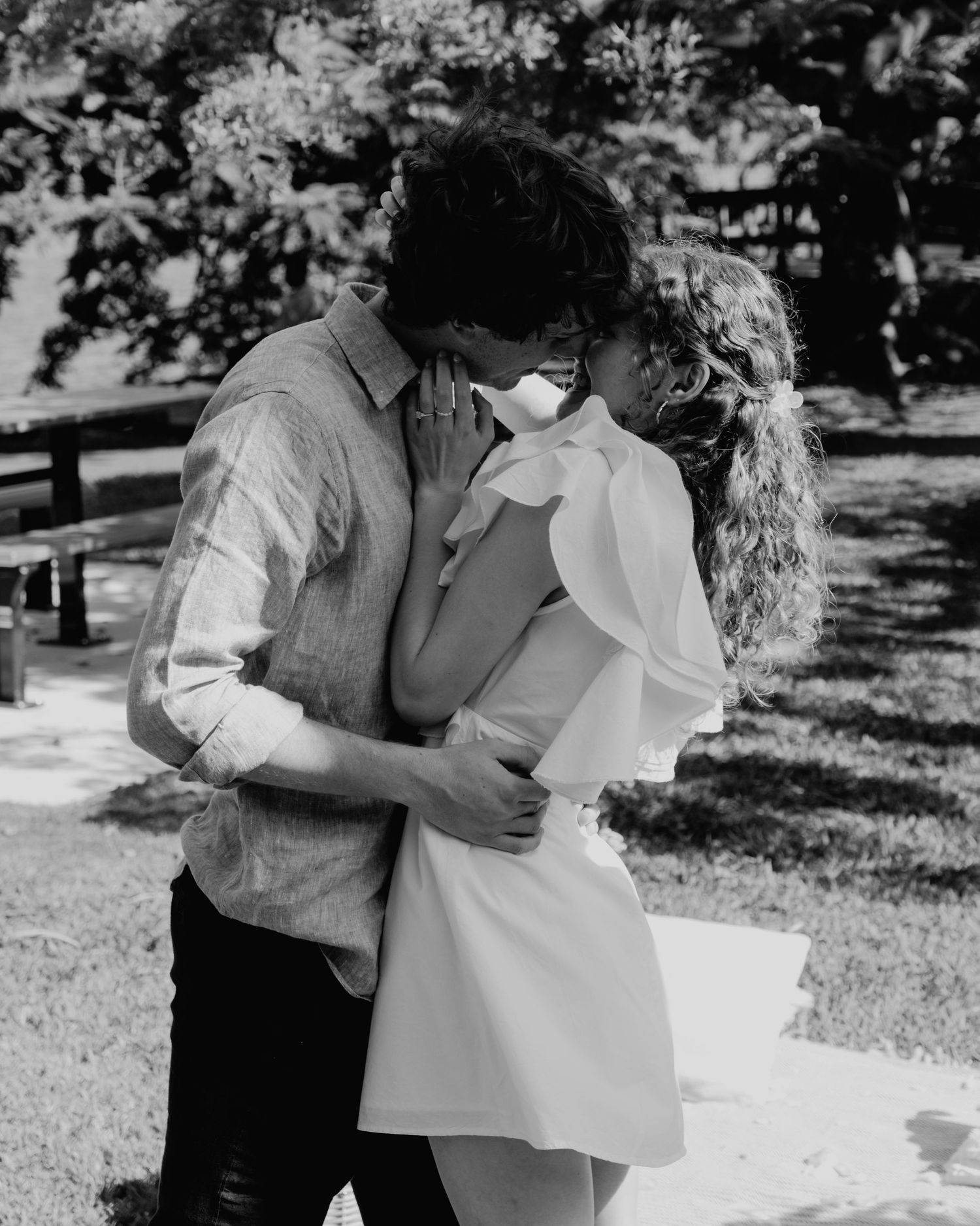 Black and white photo of a couple sharing a romantic kiss outdoors in a park setting.