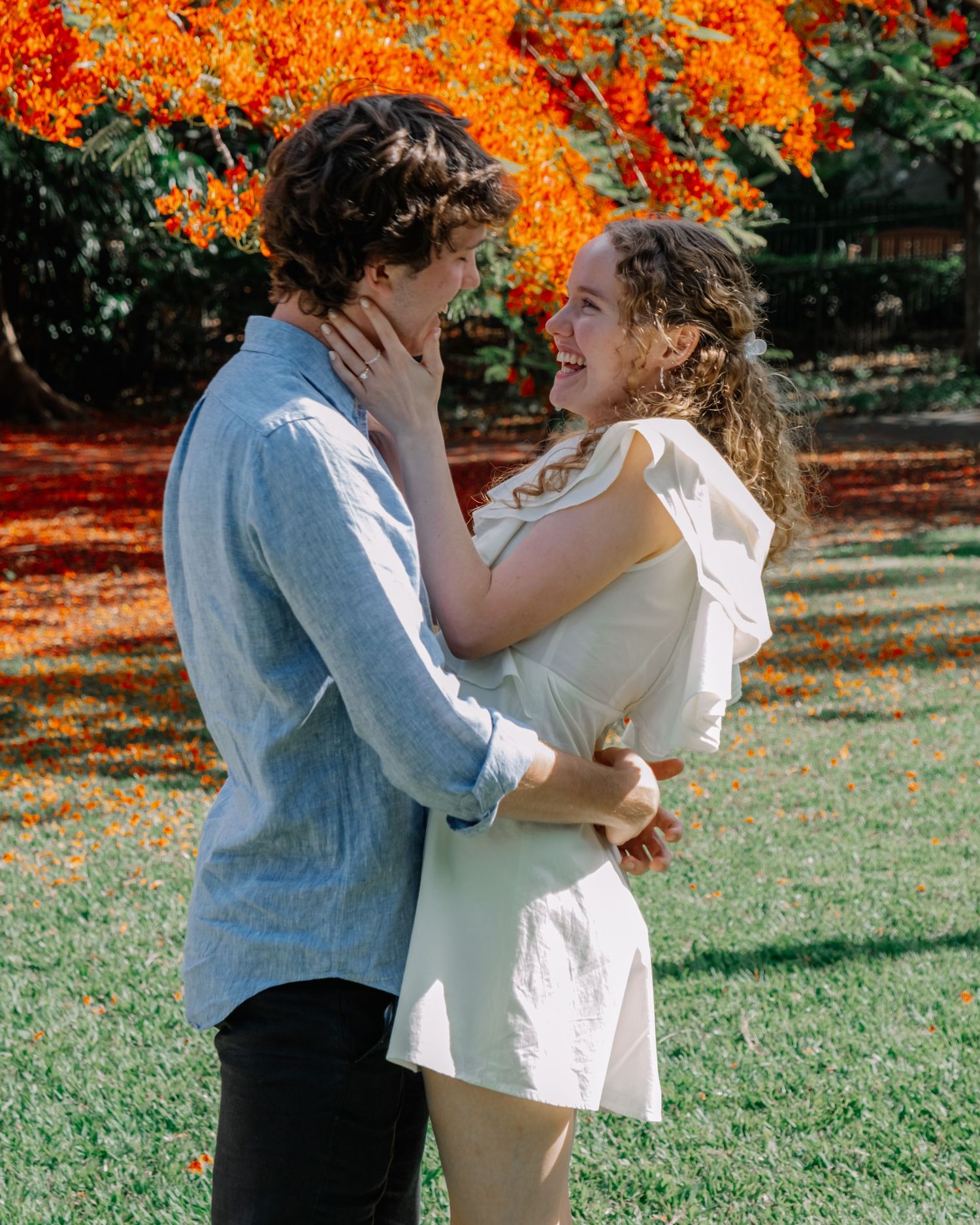 Joyful couple laughing and embracing outdoors with stunning red flamboyant tree backdrop.