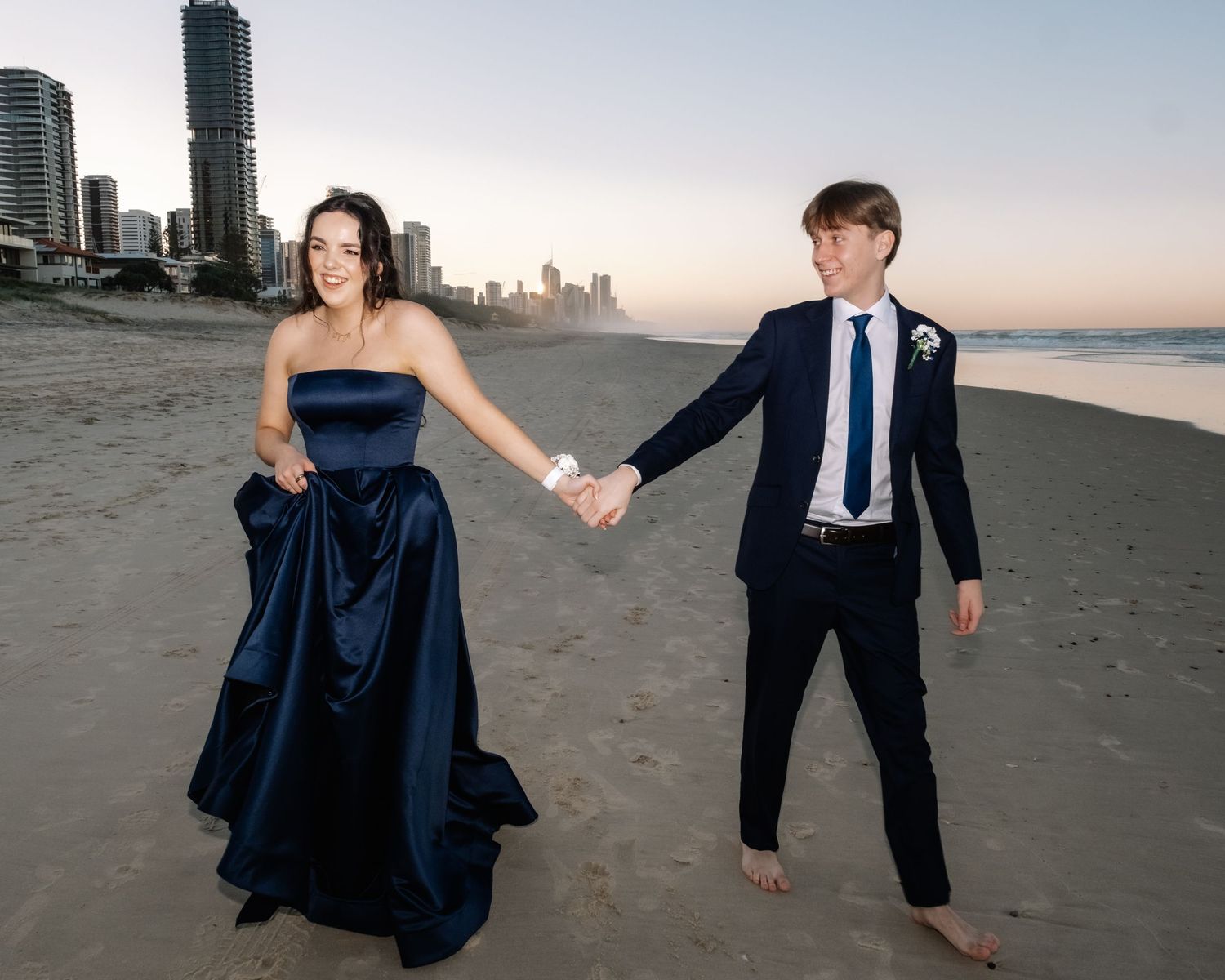 A couple in formal wear holds hands on a beach at sunset with Gold Coast skyline in the background.