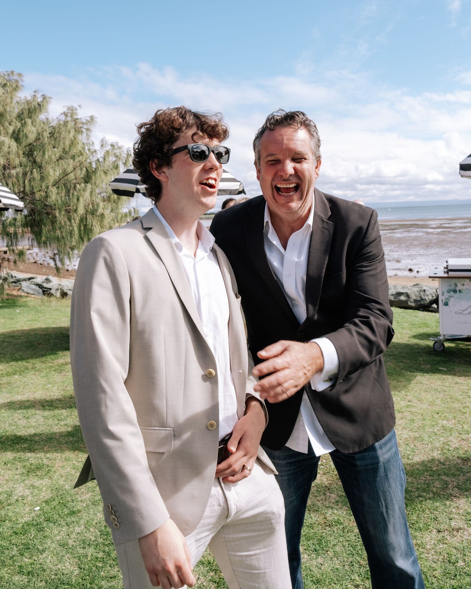 Two men laughing together outdoors at a waterfront wedding reception.