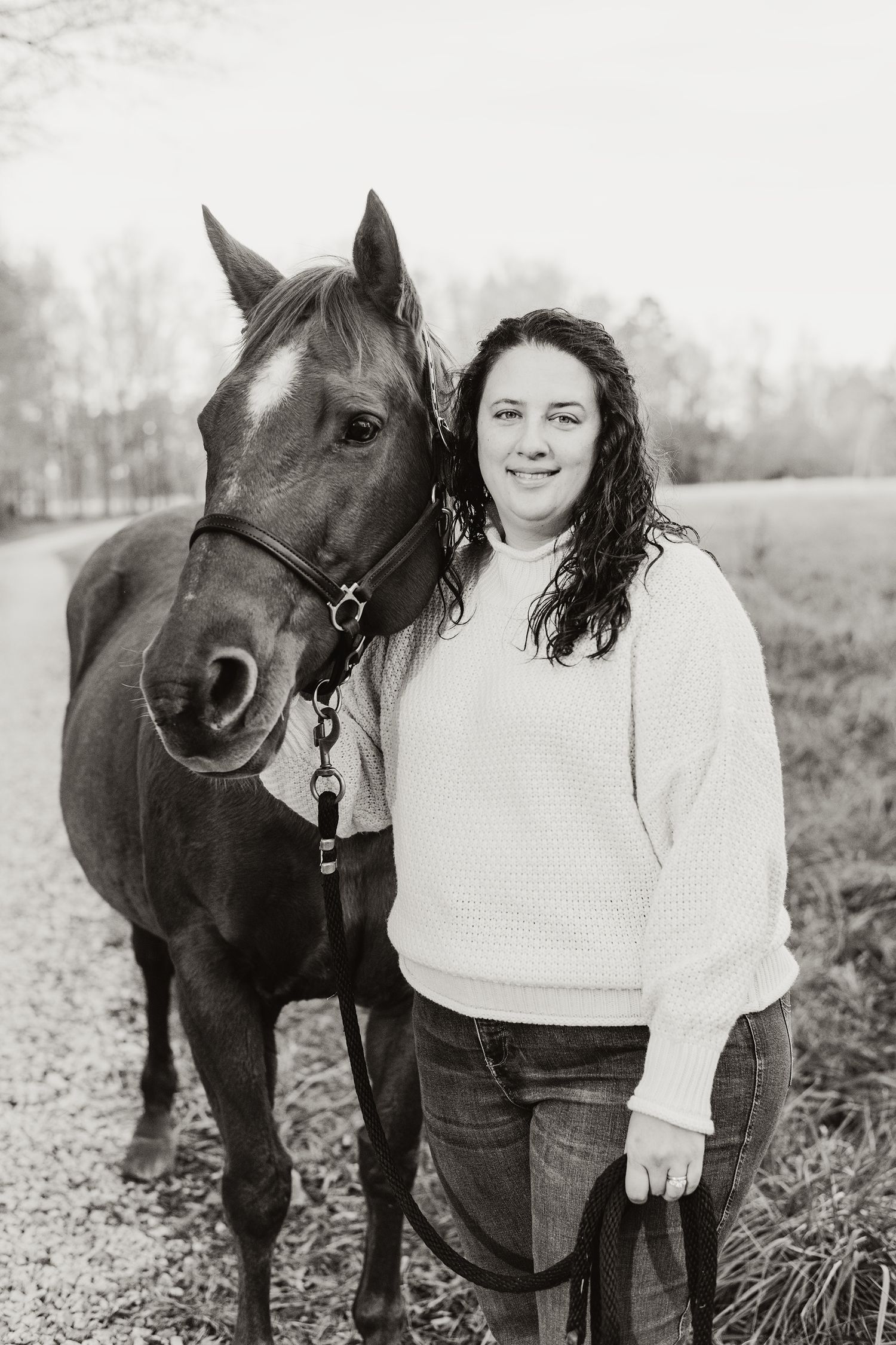 Black and white portrait of a woman standing beside her bay horse on a path.