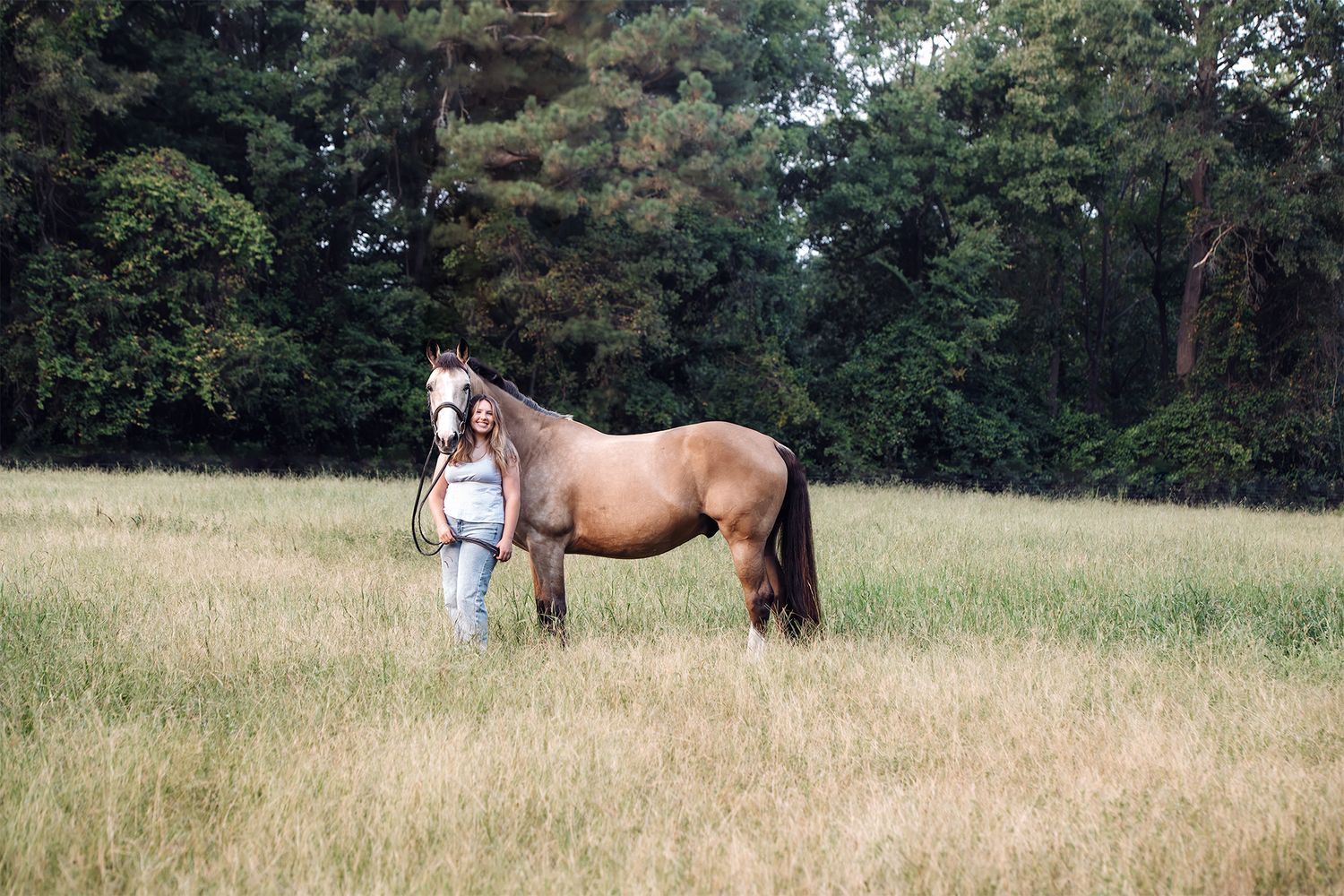 Woman standing with a buckskin horse in an open grassy field surrounded by trees.