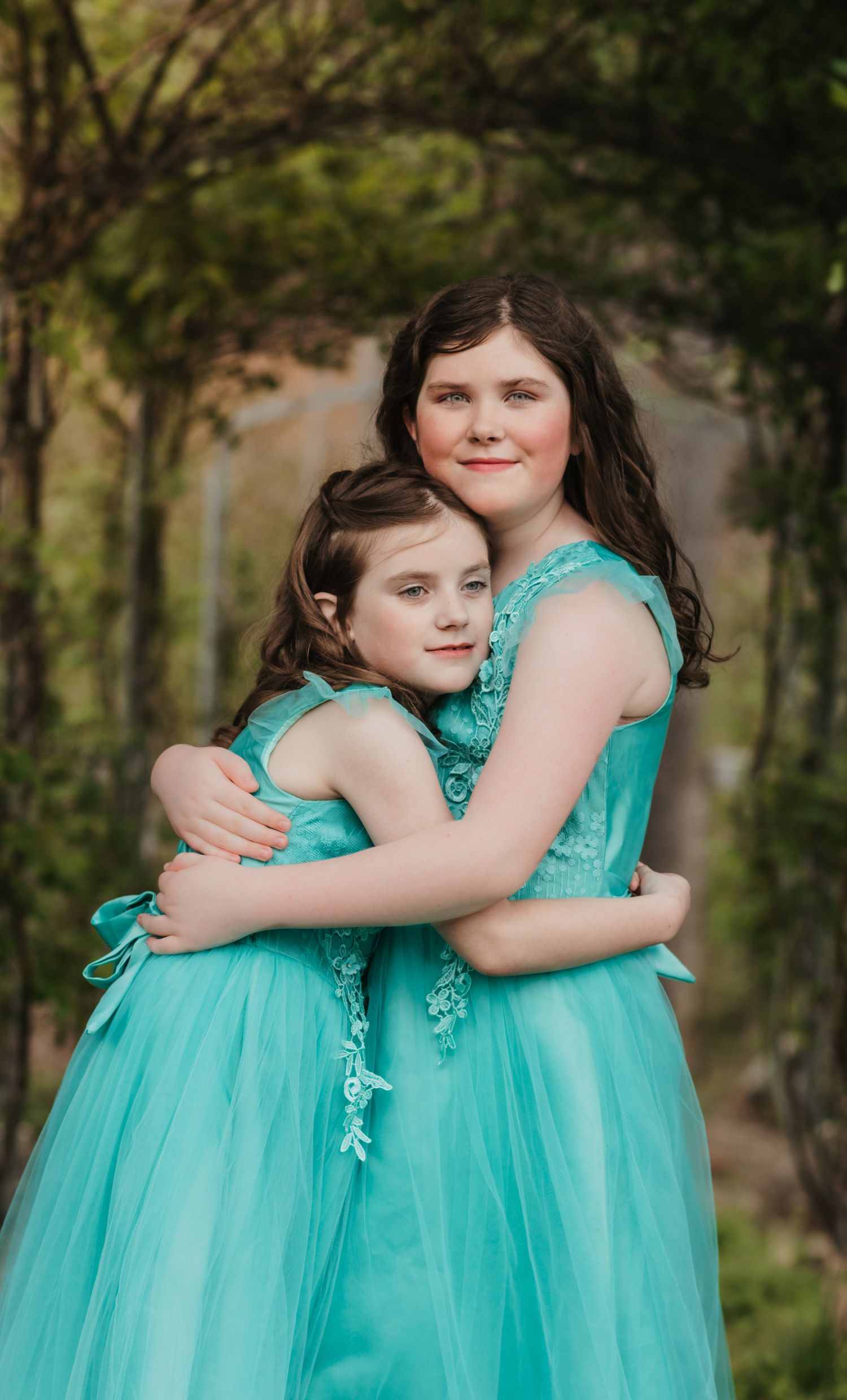 Two sisters hugging in matching teal tulle gowns under garden archway outdoors.