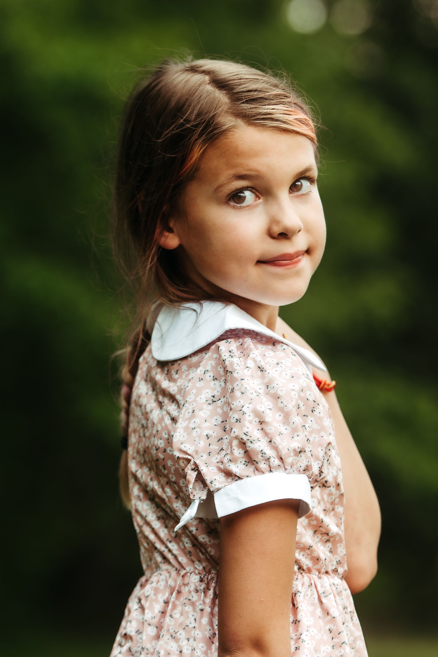 Young girl in floral dress smiling over shoulder against green bokeh background.