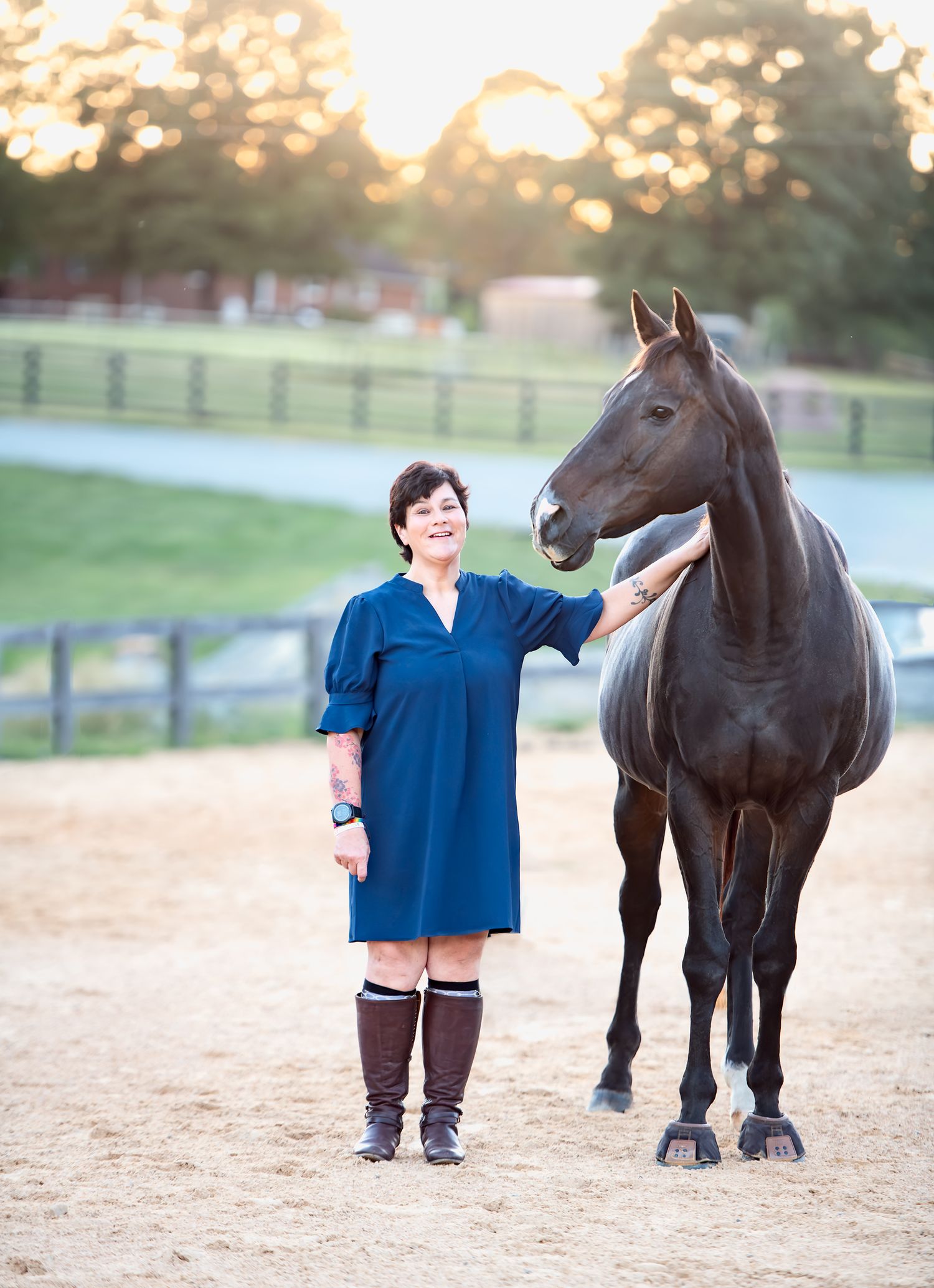 Woman in blue dress and tall boots stands smiling next to a dark horse at sunset on a farm.