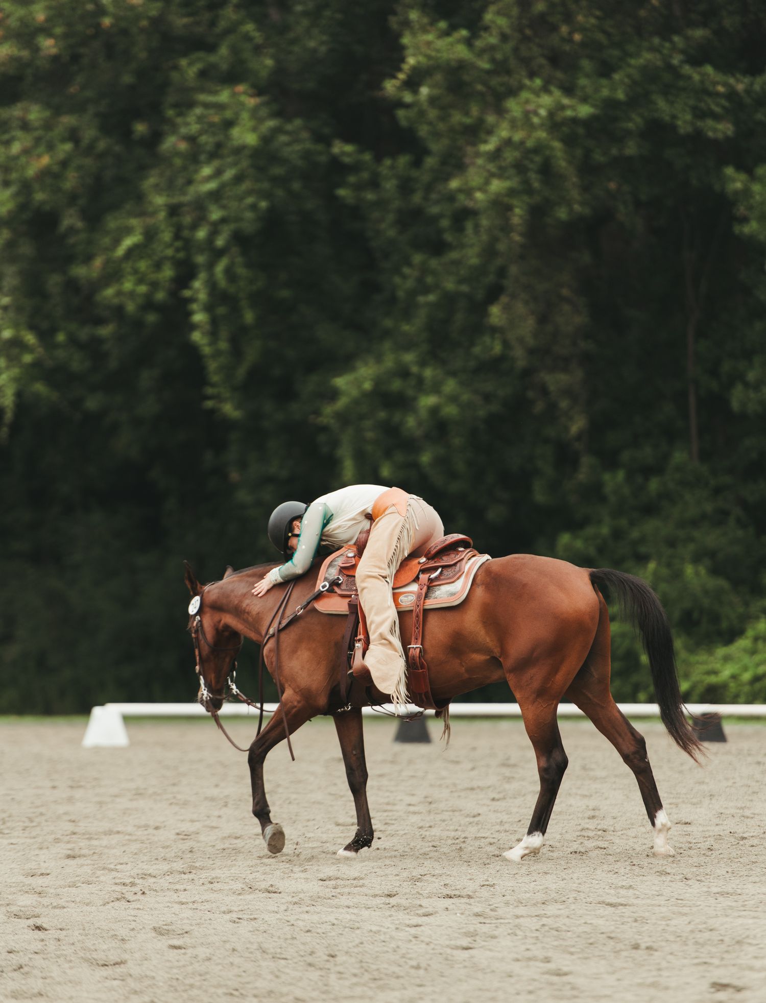 Rider leaning forward hugging a chestnut horse while riding in an outdoor sand arena.