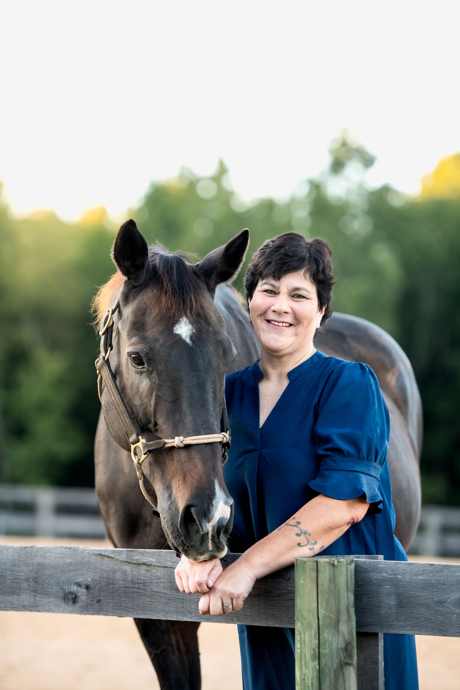 Smiling woman in blue dress posing with dark bay horse at wooden fence at sunset.