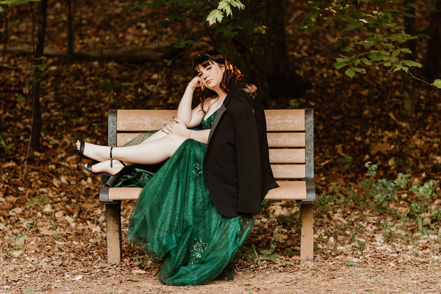 Young woman in green ball gown and black blazer resting on wooden bench in autumn forest.