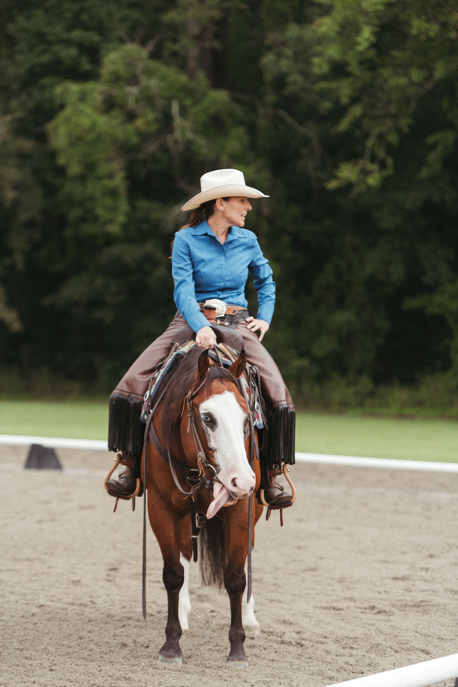 A woman in a cowboy hat and blue shirt rides a chestnut horse in an outdoor arena.