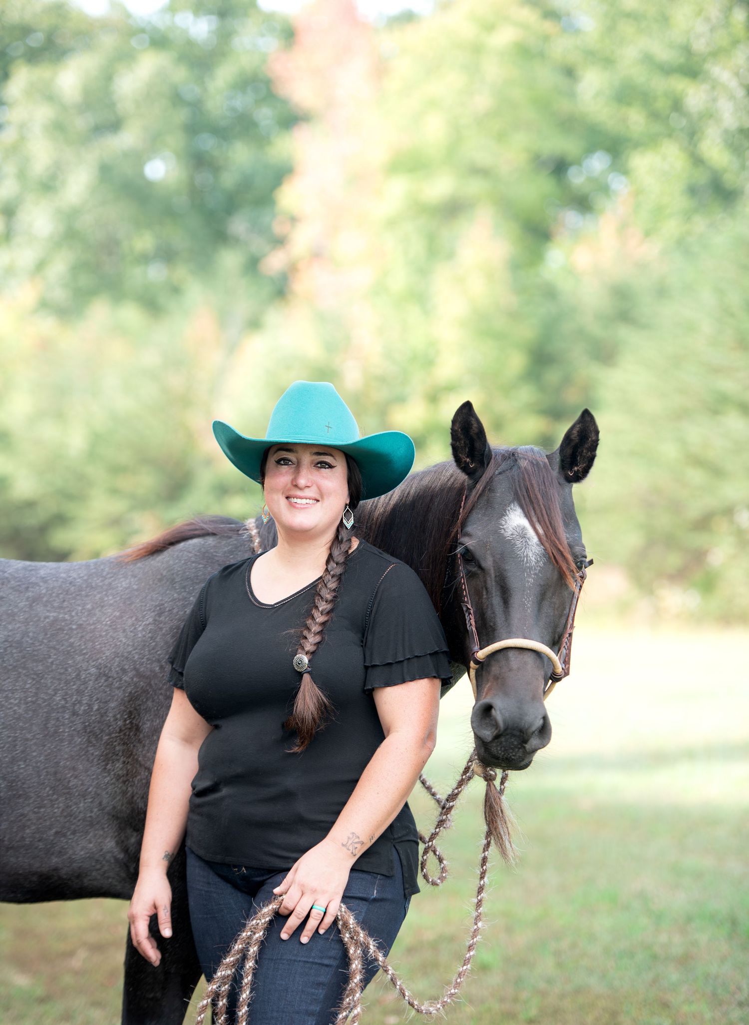 Smiling woman in a turquoise cowboy hat and black top with a long braid stands beside a dark gray horse in a sunny grassy field.