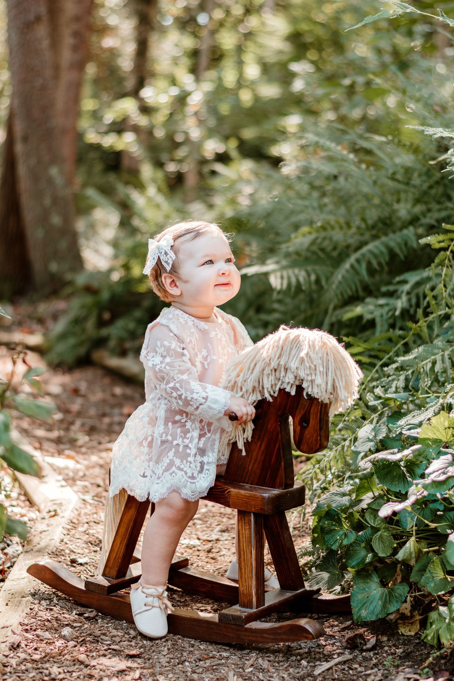 Smiling toddler in white lace dress riding a wooden rocking horse in forest.