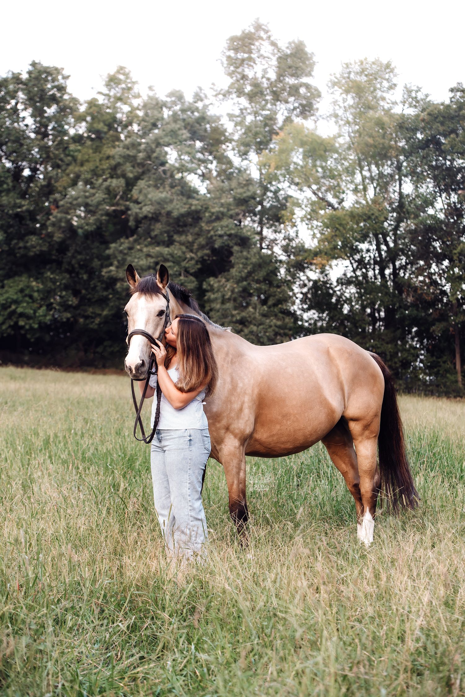 Woman in light jeans and a white top leans her face against a buckskin horse in a grassy field with trees in the background.