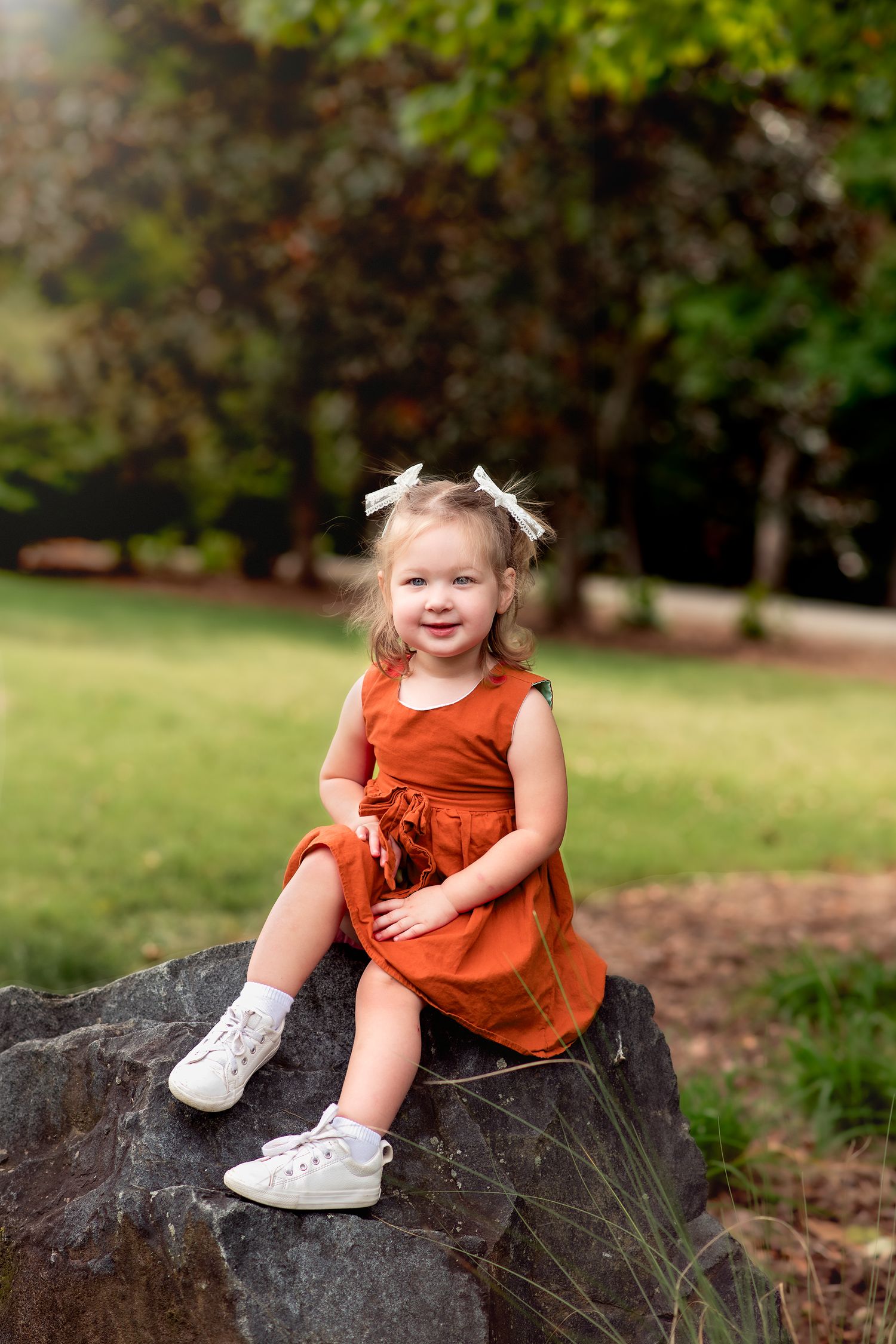 Smiling toddler girl in a rust orange dress and white hair bows sits on a large rock in a sunny green park.