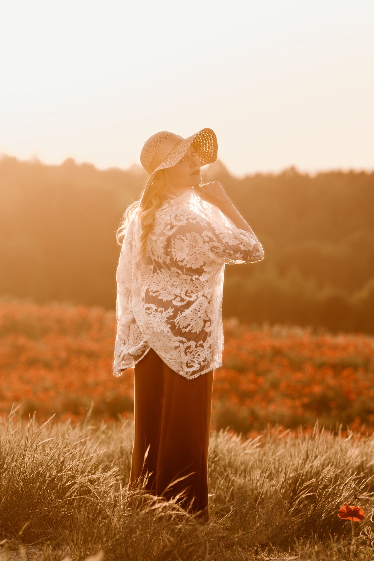 Woman in white lace top and straw hat standing in golden sunset light overlooking a red poppy field.