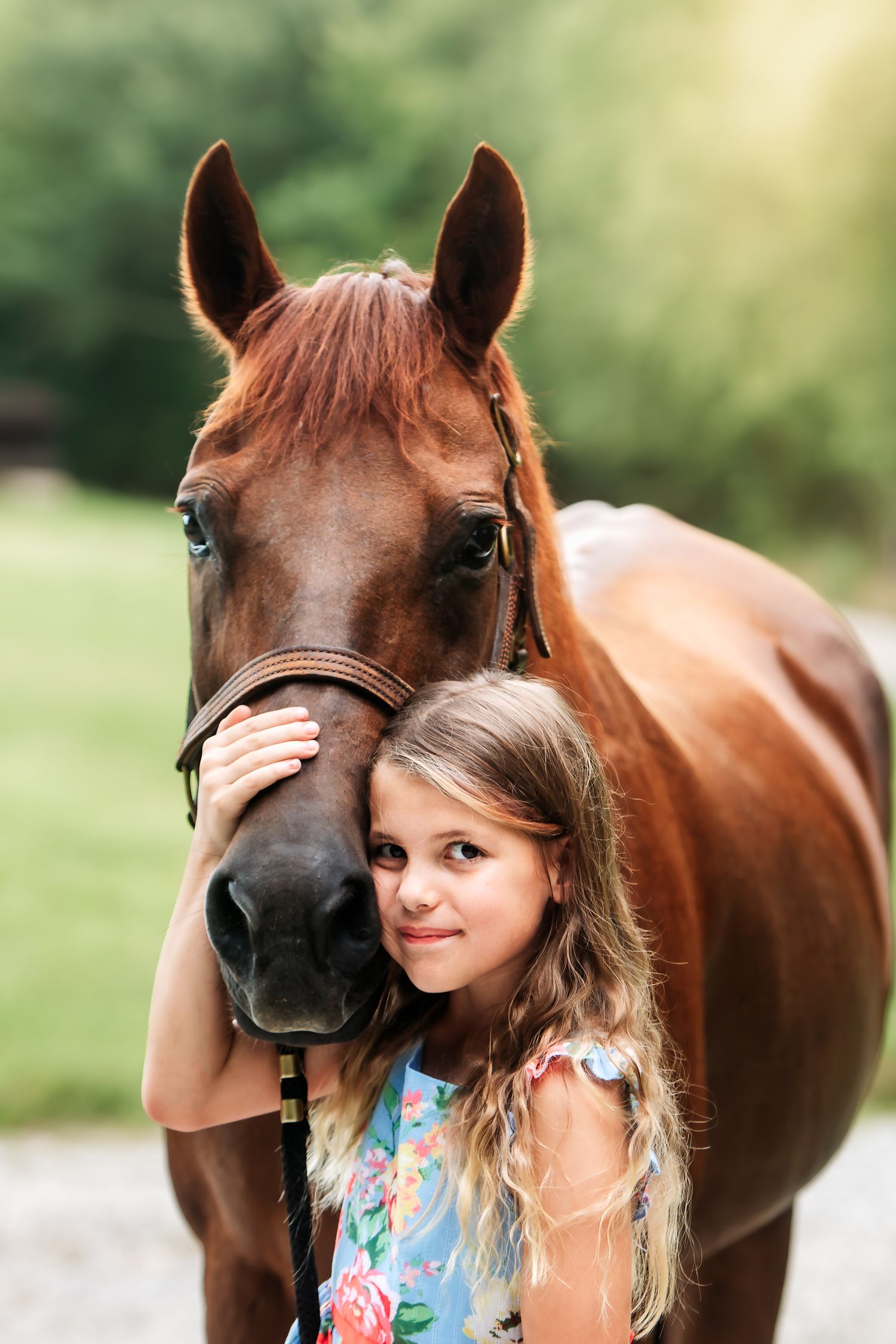 Smiling young girl in floral dress hugging a chestnut horse's nose in natural light.