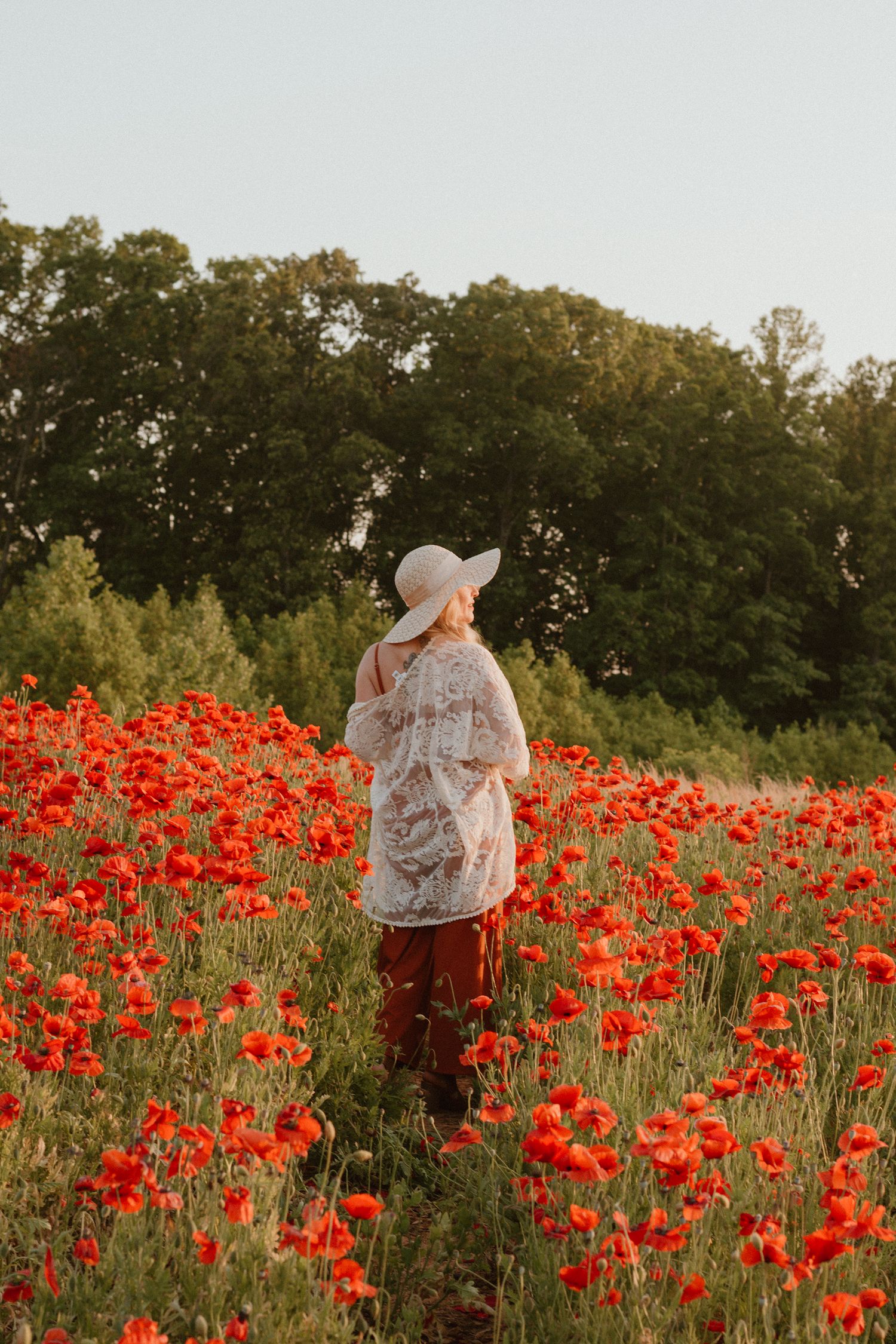 Woman in white hat and lace top walking through a vibrant red poppy field.